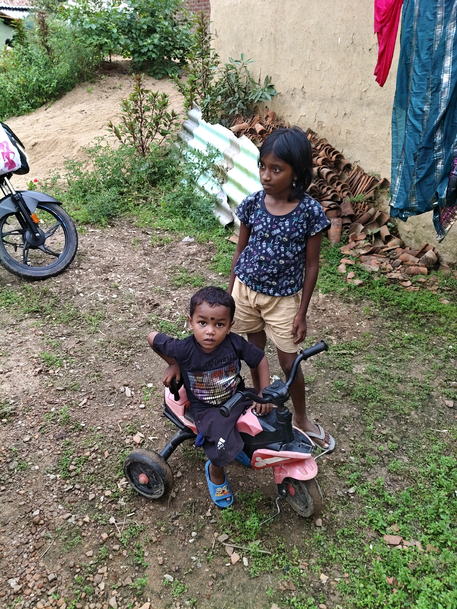 In Sosai, India, a young boy sits on a pink tricycle. He wears a black t-shirt with a colorful, patterned design and dark shorts. On his feet are blue flip-flops. Beside him, a young girl stands. She is wearing a dark t-shirt with a similar pattern and light-colored shorts. Her dark hair is pulled back. They are in an outdoor environment with a dirt and gravel ground covered in sparse green grass and small rocks. In the background, a sandy mound rises, covered with greenery. Further back, a rough-textured wall stands, with a pile of broken terracotta roof tiles leaning against it. Colorful fabrics, one pink and one blue with silver stripes, hang from above. To the left of the scene, the front wheel and fender of a black motorcycle are visible. The overall setting appears to be a humble, outdoor area, possibly a residential yard or a village roadside.