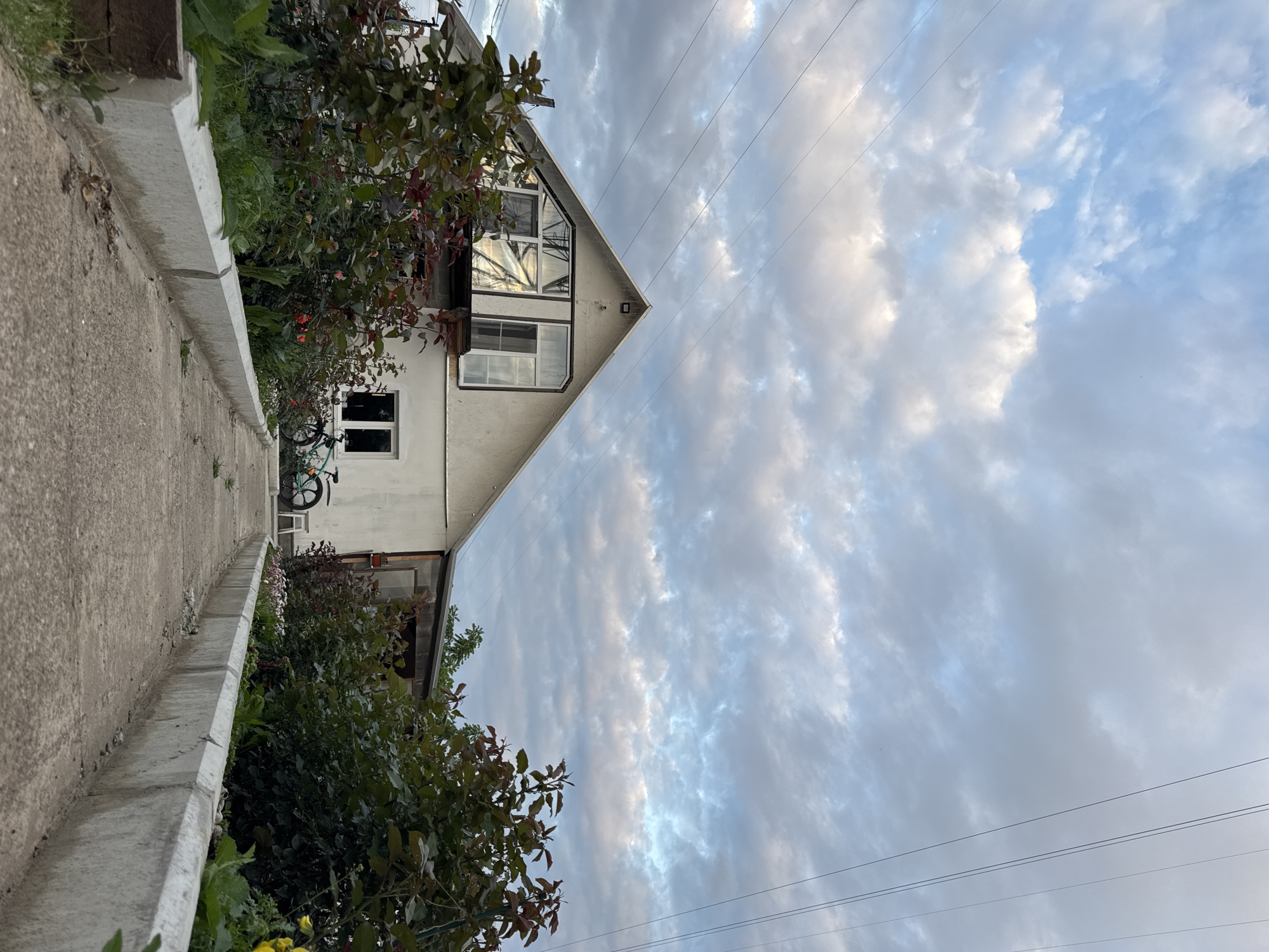 The image shows a house with a large window on the upper floor, a bicycle, and a path leading to the house. The sky is cloudy. The scene appears to be located in a residential area in Chaikovsky, Russia. The house is white with a dark roof and a porch. The bicycle is green and is parked near the house. The path is made of concrete and is lined with plants. The overall impression is of a peaceful, rural setting.