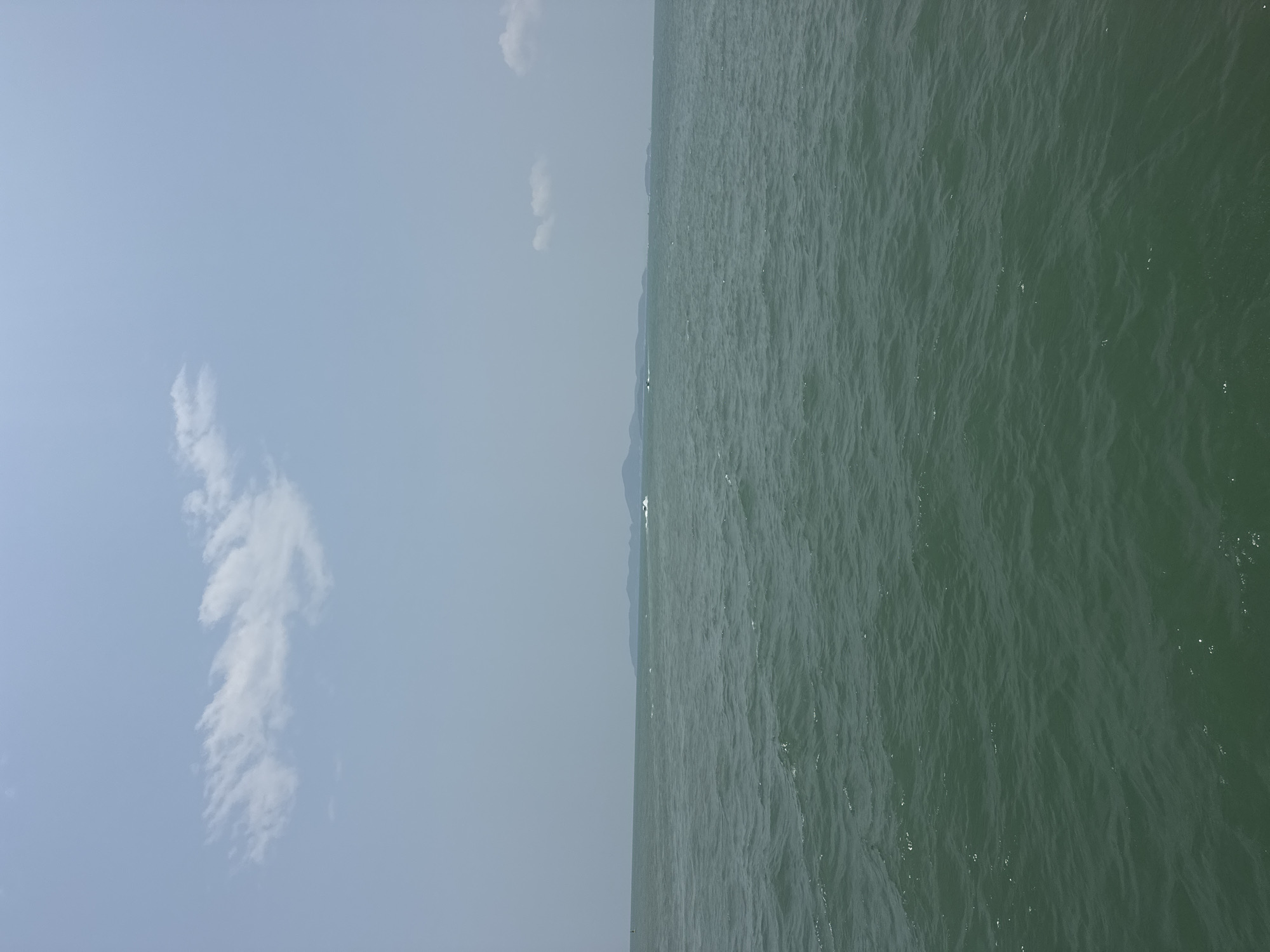 The image depicts a view of the ocean from Pattaya City, Thailand. The scene is divided into two distinct sections: the left side showcasing the sky and the right side featuring the ocean. The sky is light blue with scattered white clouds. The ocean water is a dark green color with visible wave patterns and light reflections. A landmass is faintly visible on the horizon where the sky meets the ocean.