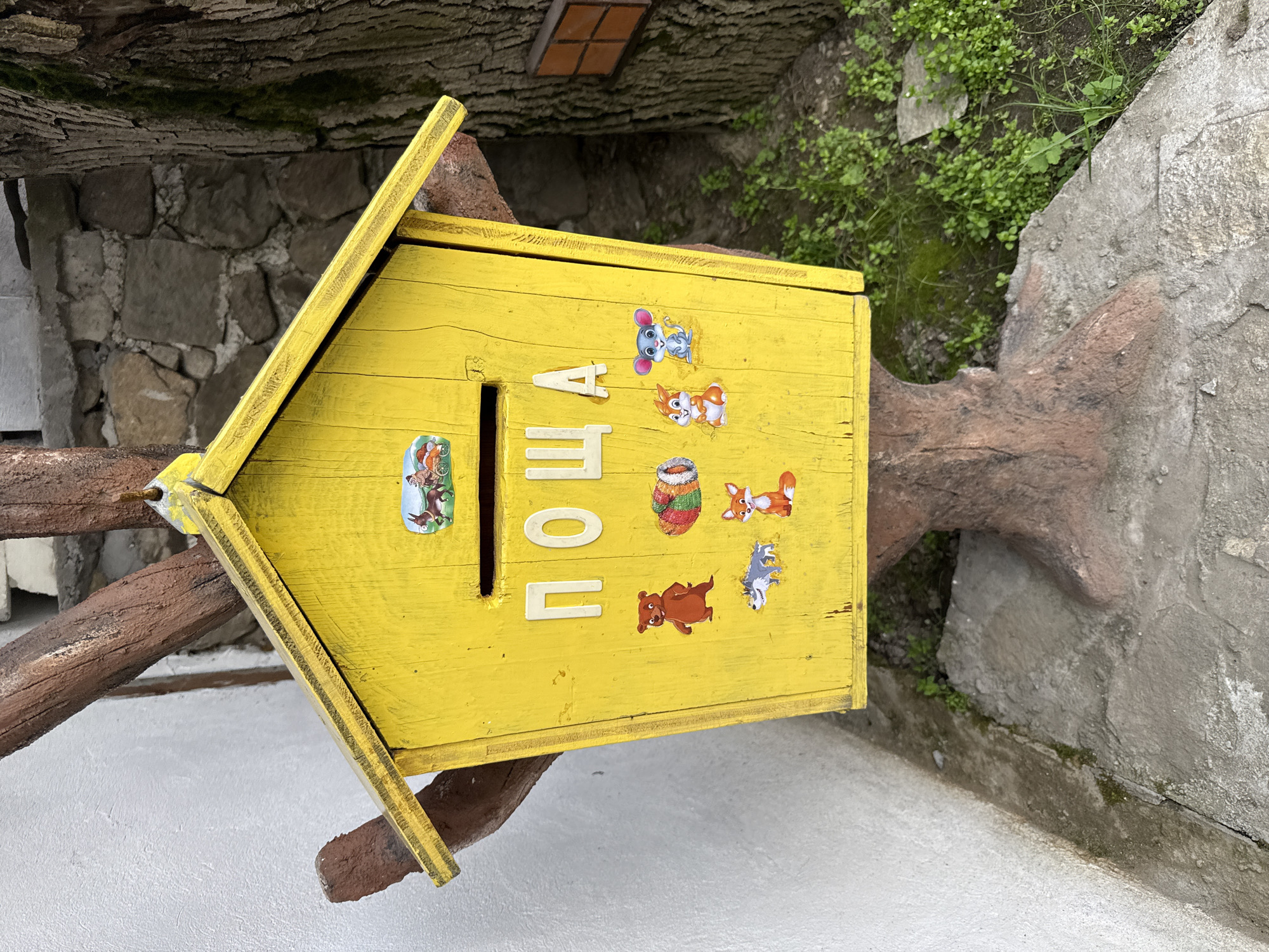 A yellow mailbox, decorated with animal stickers and the word "ПОЩА" (mail), is mounted on a brown, artificial tree branch. The background consists of stone walls, some greenery, and what appears to be a tree trunk. The mailbox is located in Zlatitsa, Bulgaria.