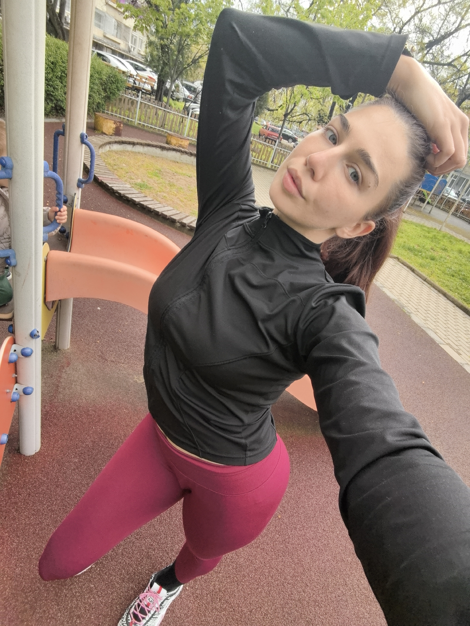 This self-portrait captures a young woman in an outdoor playground setting, likely in Stara Zagora, Bulgaria. She is the central figure, looking directly at the camera with a neutral expression. Her dark hair is pulled back into a ponytail, and she wears a black long-sleeved athletic top and dark pink/magenta leggings, paired with athletic shoes featuring pink laces and black socks. A partial "NIKE" logo is visible on her shoe, along with what appears to be "ALI" text below it. Her right arm is extended upwards, holding the camera for the selfie, while her left arm is bent with her hand resting in her hair.
The background reveals typical playground equipment, including a light-colored structure with blue handles and an orange slide. To the left, a child's hands are visible holding onto the blue handles, indicating another person is present on the playground. The ground is covered with a reddish-brown rubberized safety surface. Beyond the playground, there's a grassy area, a wooden fence, parked cars, and residential buildings, suggesting an urban park environment. The trees, with nascent green leaves, and the overcast sky point to an early spring day with diffused lighting. The overall scene depicts a casual moment outdoors, possibly during a visit to the park.