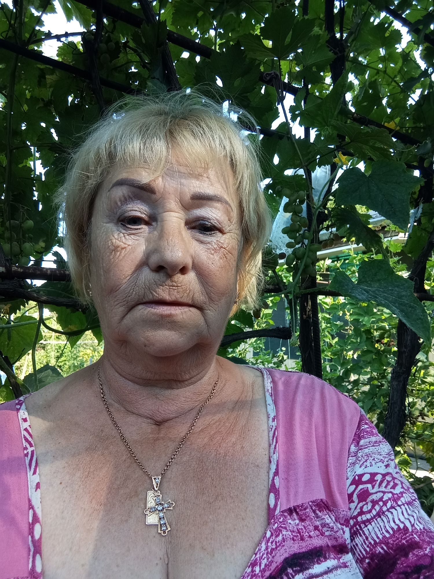 A middle-aged woman with blonde hair and wrinkled skin is pictured outdoors. She is wearing a pink top with a white and purple pattern and a gold chain with a cross pendant. The woman is positioned in the foreground, with a backdrop of green foliage, likely a grapevine, and a glimpse of a building and other vegetation in the distance. The scene appears to be in a garden or yard.