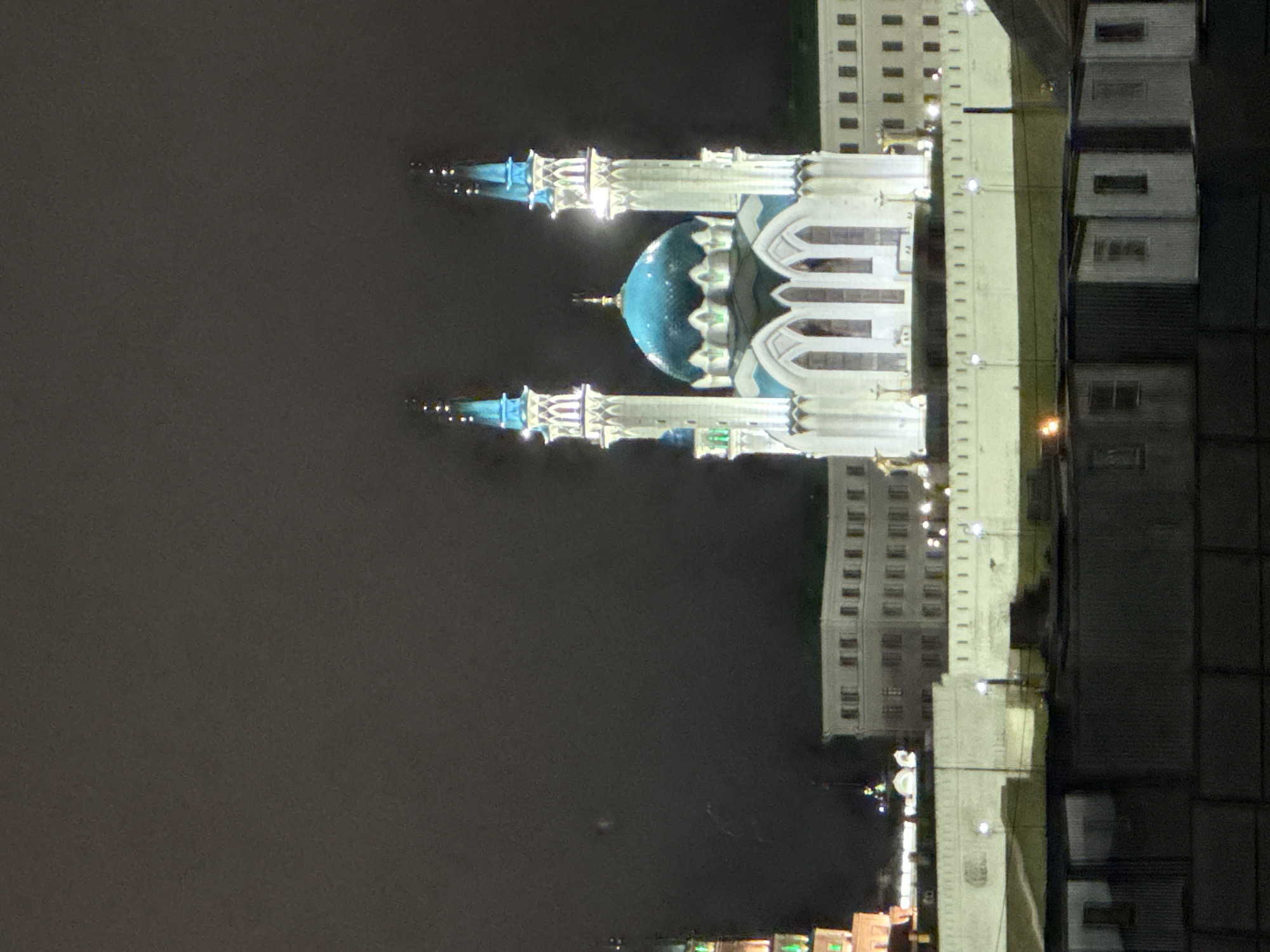 The image shows the Qolsharif Mosque in Kazan, Russia, reflected in water at night. The mosque has a blue dome and white minarets, illuminated against a dark sky. Adjacent buildings with lit windows are visible, also reflected in the water.