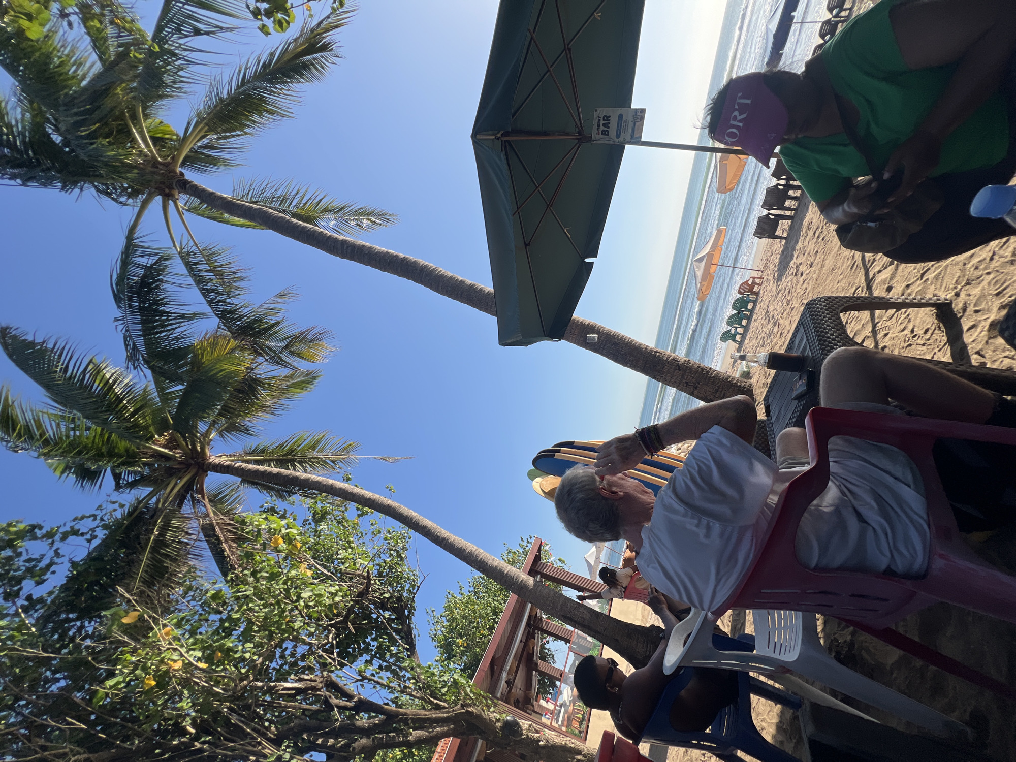 This image captures a scene on Kuta Beach, Indonesia, under a clear blue sky. In the foreground, two individuals are seated at a table on the sand. One person, with gray hair and wearing a white t-shirt, is looking towards the right, resting their hand on their forehead. The other person, wearing a green shirt and a pink visor with "PORT" visible, is looking down at something in their hands. Behind them, surfboards are propped up.
The background features the ocean with gentle waves, and several beach umbrellas and chairs are scattered across the sandy shore. Palm trees with lush green fronds are visible, casting shadows on the beach. A building with a red roof and open-air seating is situated to the left, with more trees surrounding it. A sign is attached to the umbrella pole in the middle ground, with text partially obscured but readable as "GERRY BAR." The overall atmosphere suggests a relaxed setting at a beachside establishment.