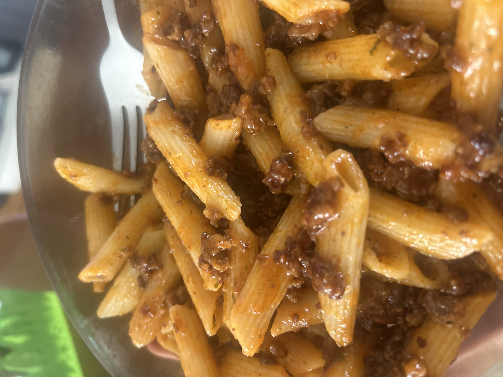 The image shows a close-up of a bowl of penne pasta with a meat-based sauce. A white fork is partially visible in the upper left corner. The pasta and sauce appear glossy. The background is out of focus, but some green and pink colors are noticeable. The location is not discernible from the image, so Burgas, Bulgaria, cannot be confirmed.