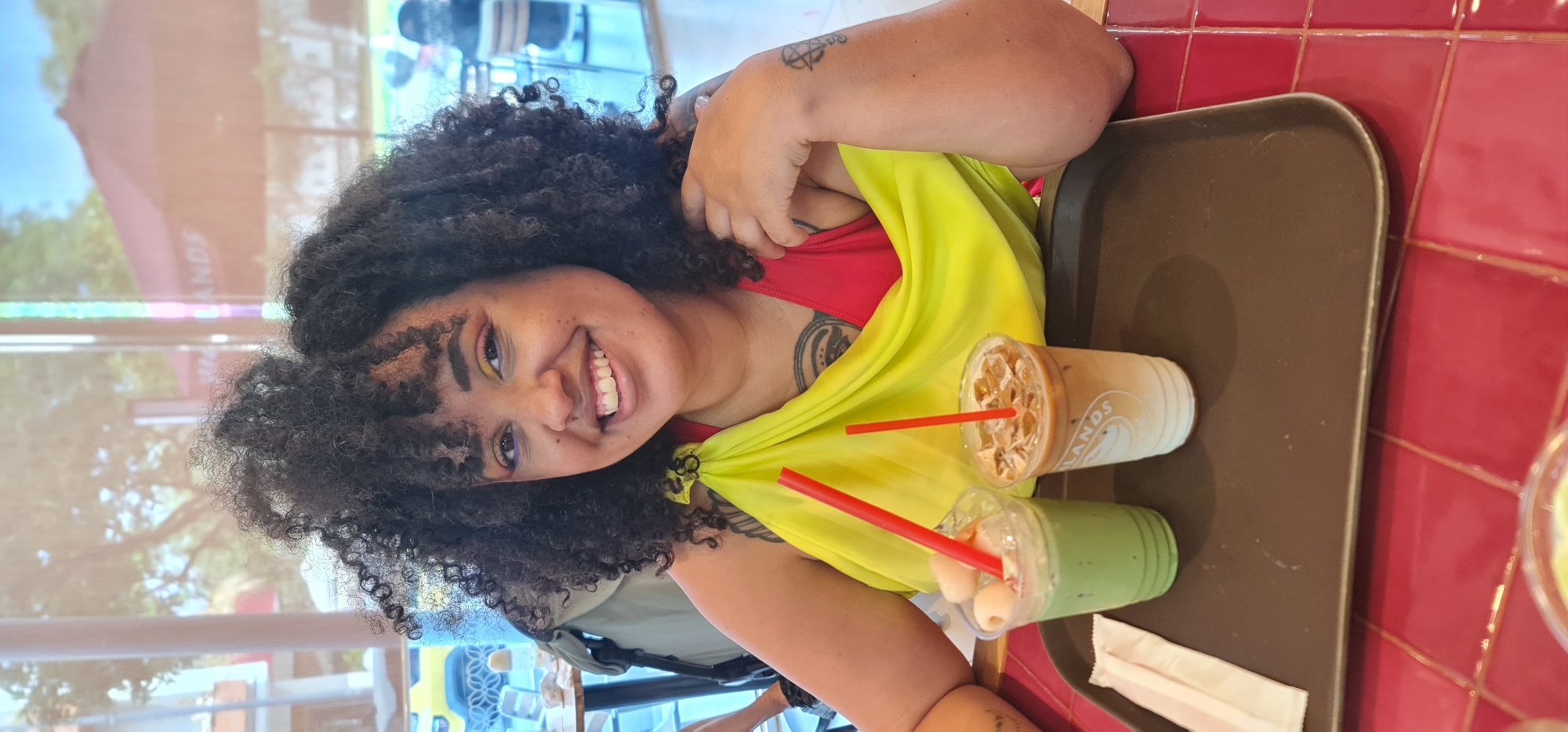 A woman with curly hair is photographed indoors at a location in Đà Nẵng, Vietnam. She is smiling, wearing a red top and a yellow shawl. A tray holding two drinks with red straws sits next to her on a table. One drink is brown with ice and appears to be coffee-based; the other is green and contains small white spherical objects. The background is blurred but suggests a cafe or similar establishment with outdoor views.