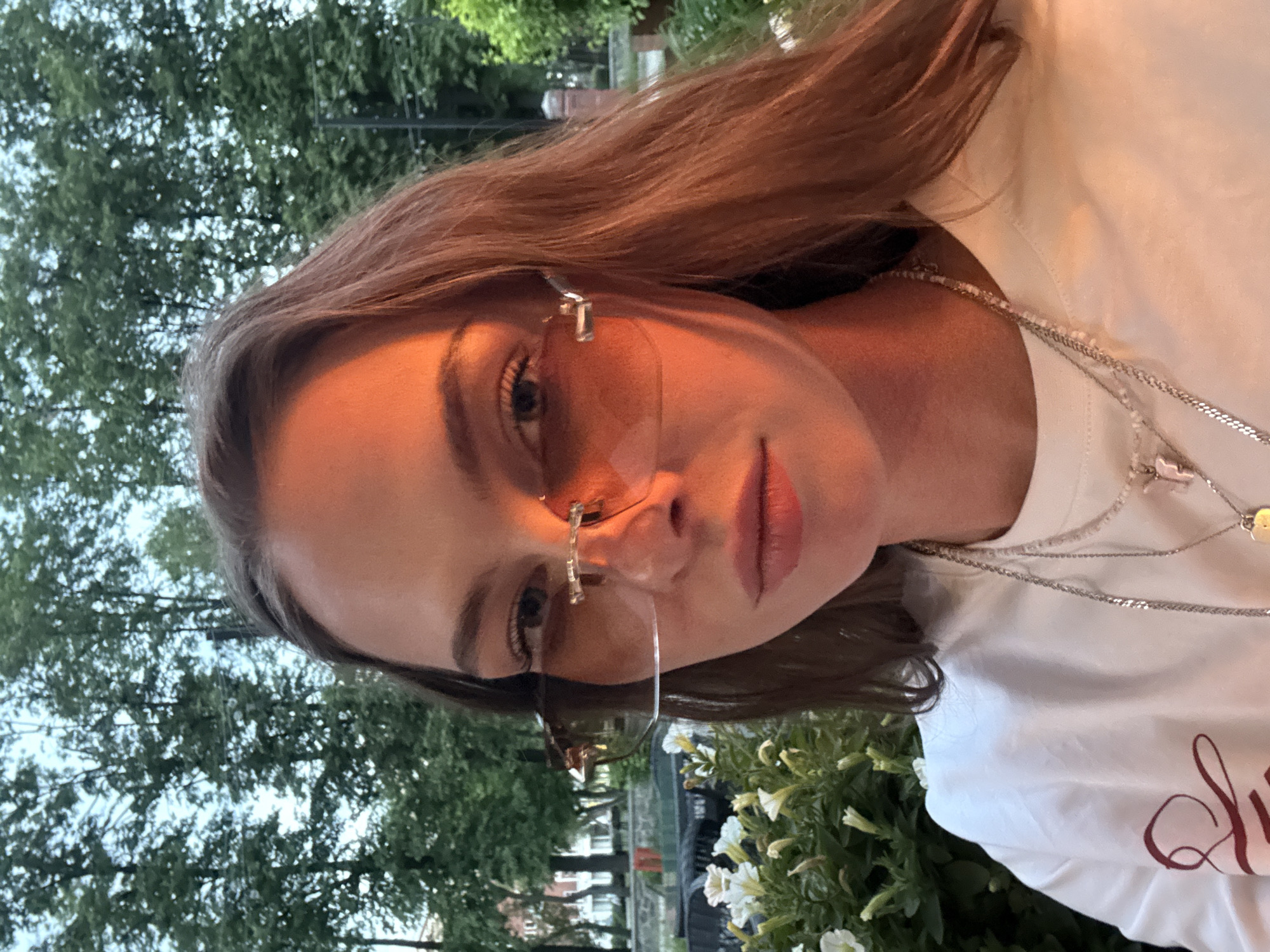 The image displays an upper-body portrait of a young adult female outdoors in Perm, Russia. Her long, brown hair is parted on the right and falls around her shoulders. She wears rimless, aviator-style sunglasses with orange-tinted lenses and silver-colored hardware, looking directly at the camera. A strong, warm orange-red light source illuminates the right side of her face, neck, and hair, creating pronounced highlights and reflections on the sunglass lenses. She is dressed in a white crew-neck t-shirt, on the left side of which a partial red, calligraphic design is visible. Several layered necklaces adorn her neck, including a thin silver chain with a small heart-shaped pendant, a beaded necklace with a light-colored charm shaped like the letter 'F', and a thicker silver chain. In the lower left foreground, a planter containing white petunia-like flowers and green foliage is visible. The background consists of numerous deciduous trees with dense green canopies, silhouetted against a light blue, possibly overcast, sky. Through the trees, distant urban buildings, dark outdoor seating, and a brick pillar are discernible.