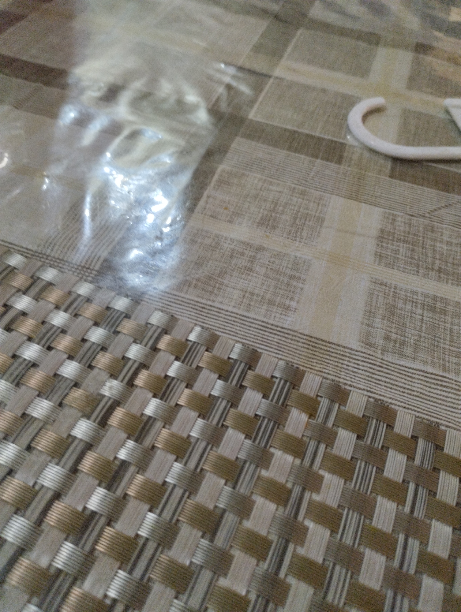 A close-up shot captures a patterned placemat and tablecloth. The placemat in the foreground is woven with alternating strips of beige, brown, and silver threads, creating a textured geometric pattern. The tablecloth beneath it has a checkered design with intersecting lines of beige and grey. A clear plastic wrap is visible in the upper left corner of the image, covering an indistinct object. In the upper right, a portion of a white curved object, possibly a hanger, rests on the tablecloth. The scene is indoors, likely on a flat surface like a table.