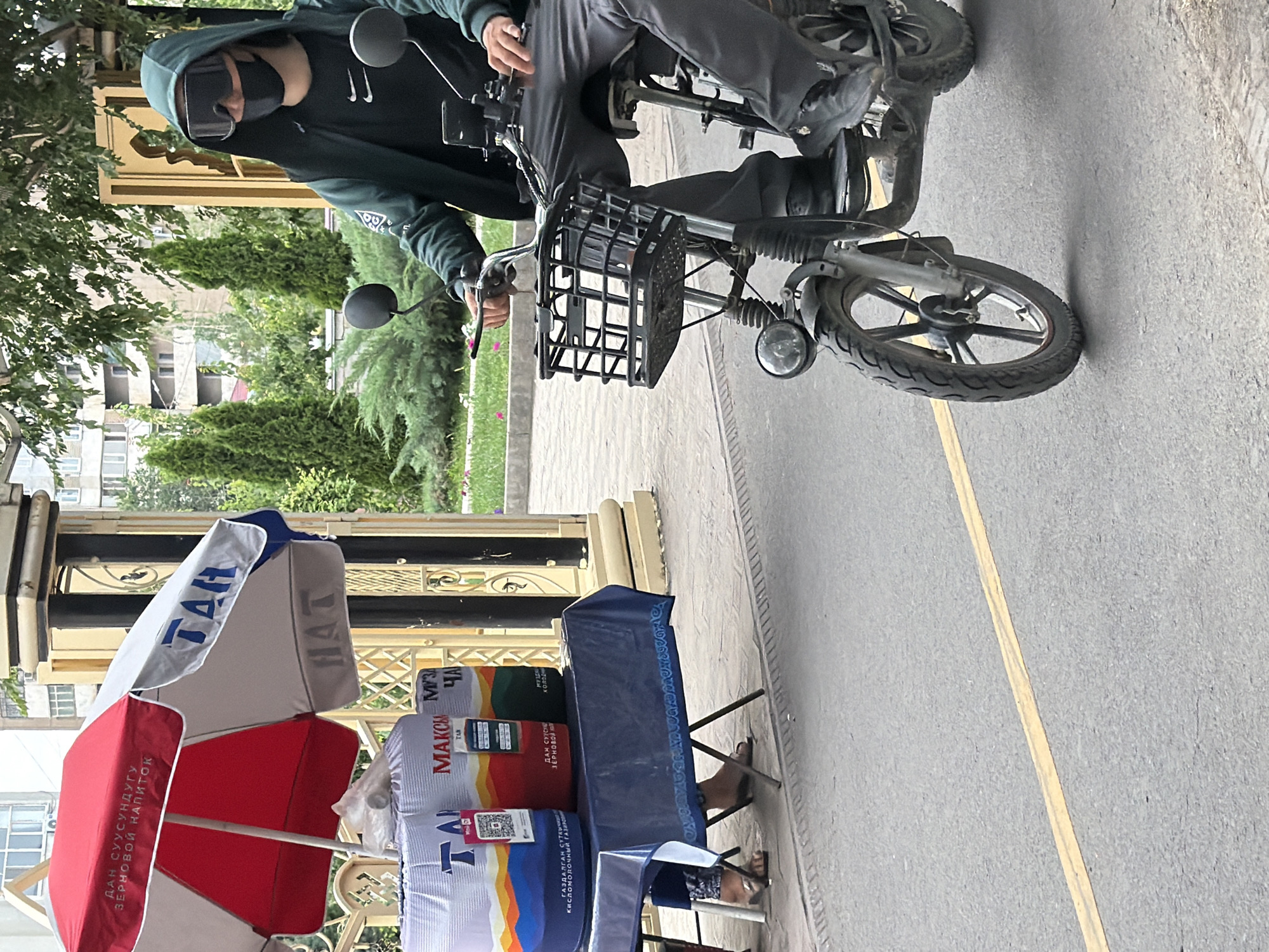 In an urban setting, a person wearing a dark hooded jacket, a black face mask, and large goggles sits on a black electric scooter. The scooter has a black wire basket attached to the handlebars. The person is holding a cell phone in their right hand. To the left of the scooter, a blue and white striped umbrella is set up over a table covered with a dark blue tablecloth. On the table are several large cylindrical objects wrapped in blue and white packaging, some with colorful designs featuring abstract shapes and landscapes. Text in Cyrillic is visible on the packaging. A person's legs and feet, wearing flip-flops, are visible under the table. Behind the table and umbrella, a decorative metal fence and a textured column are visible. The ground is paved with asphalt, marked by a yellow line. In the background, lush green trees and shrubs, as well as a multi-story building, can be seen.