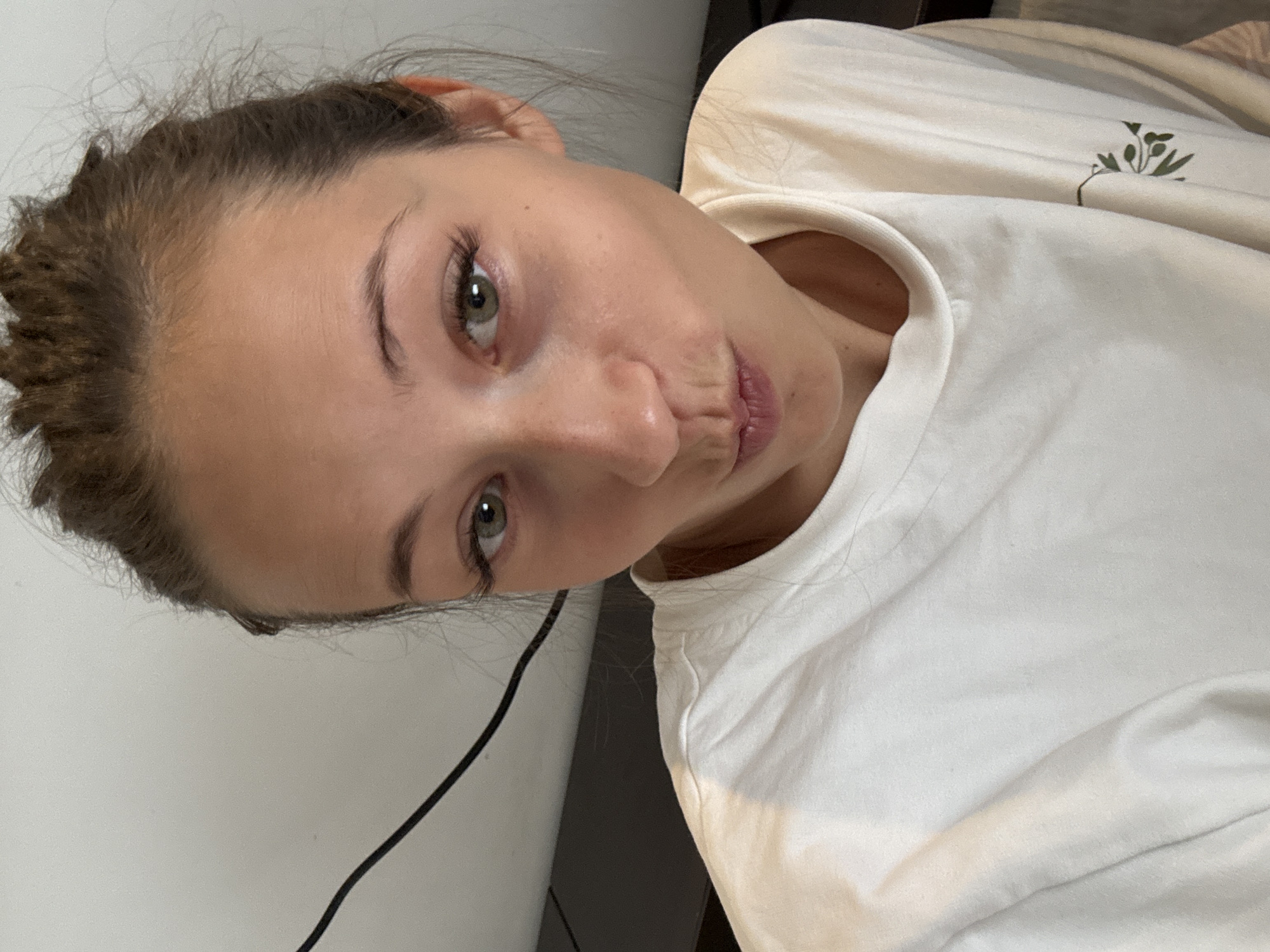A close-up, angled shot of a young person with fair skin, light brown hair pulled back, and green eyes. The person is wearing a white T-shirt with a small green plant design on the shoulder. A black cord is visible near the left edge of the frame against a white background, possibly a wall. The setting is indeterminate, with no clear indication of location, and no specific evidence to link it to Saint Petersburg, Russia.