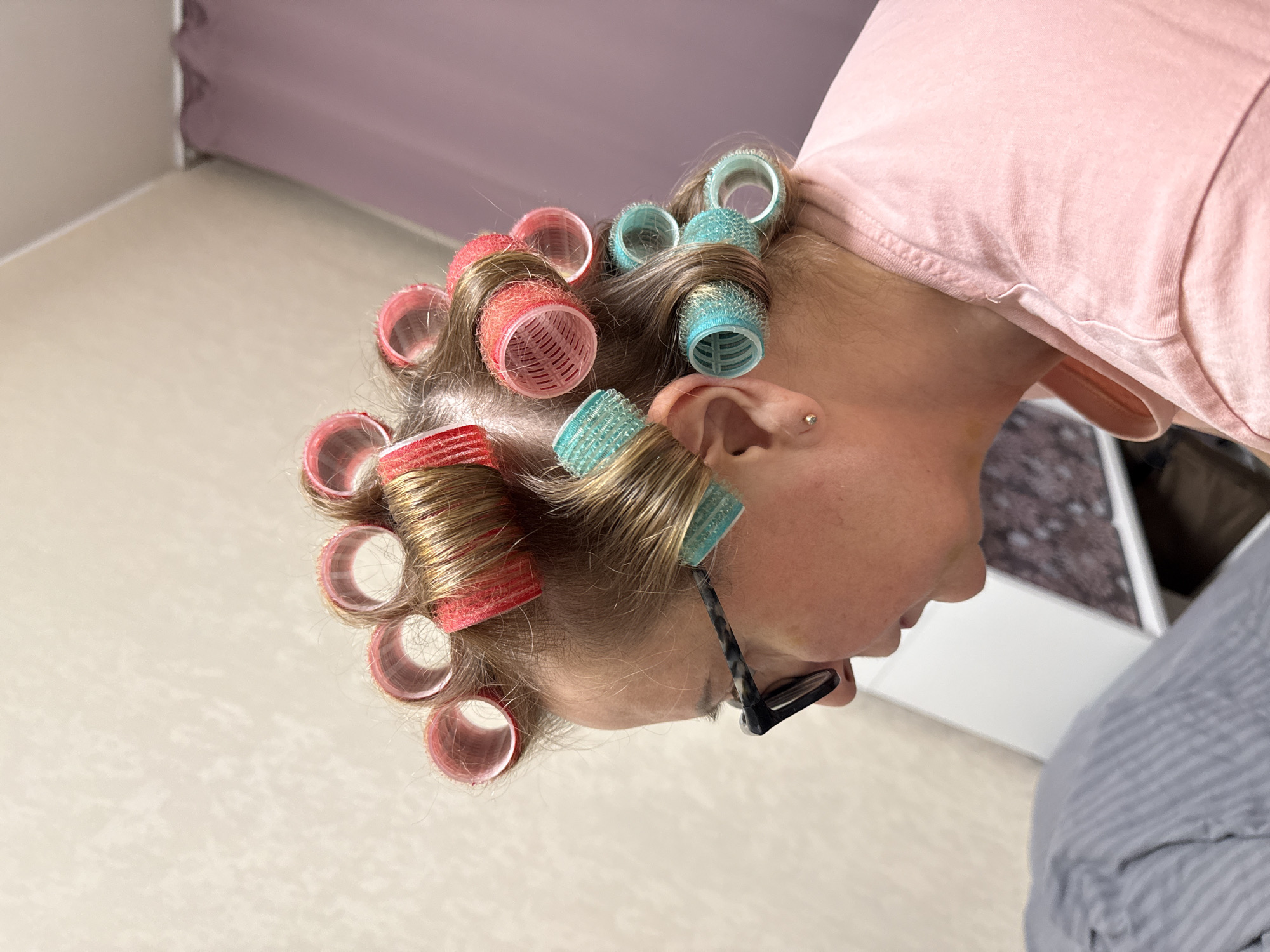 A person with light brown hair is seen wearing pink and teal hair rollers. The person also wears glasses and a light pink shirt. The background consists of a light-colored wall and a glimpse of what appears to be a striped garment and a partially visible object. There is no visible information indicating this photograph was taken in Moscow, Russia.