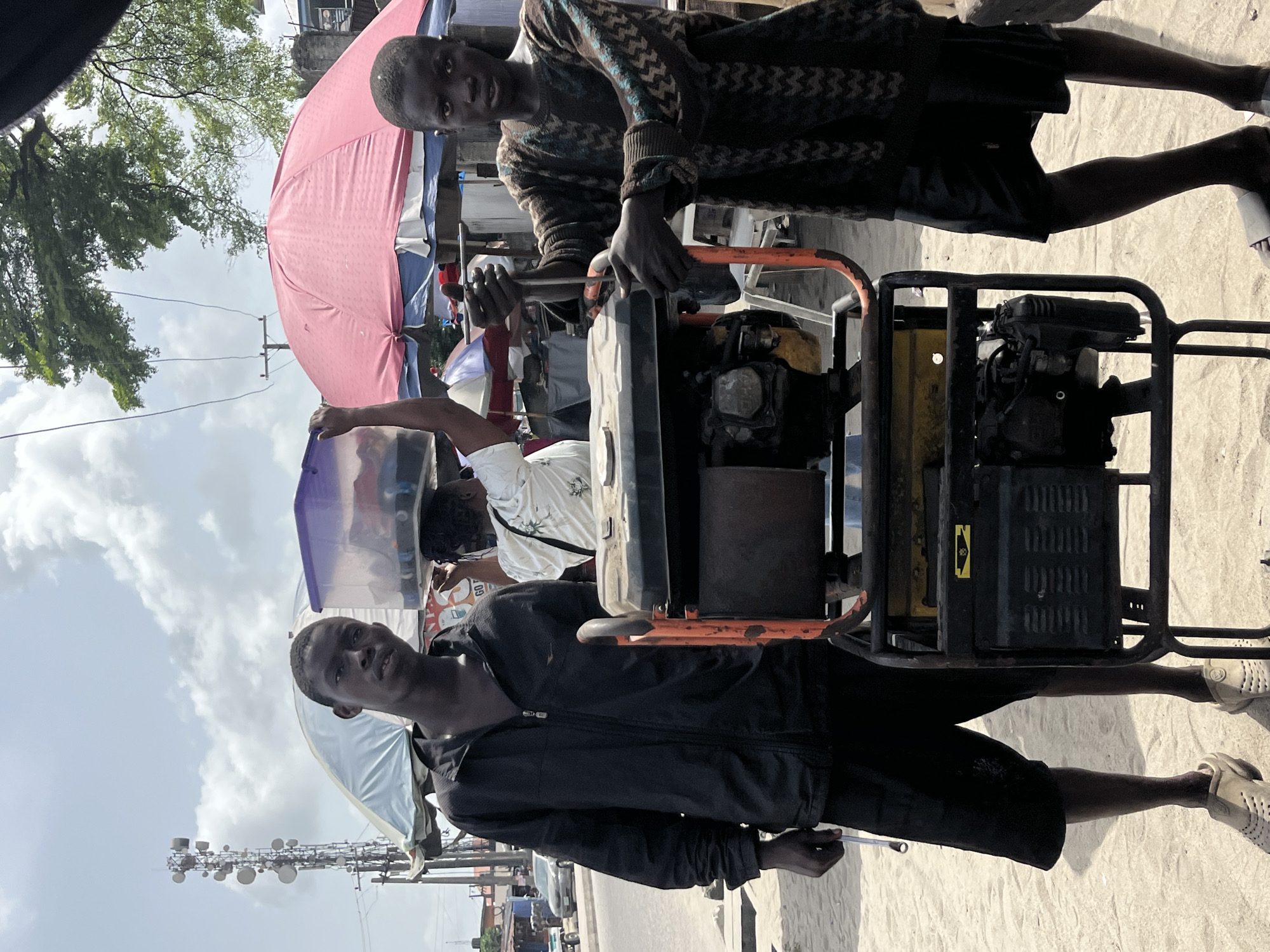 The image, taken in Ado, Nigeria, captures an outdoor scene featuring two young men and a portable generator. In the foreground, a young man wearing a patterned long-sleeved sweater and dark shorts stands beside the generator, his hands on its frame, suggesting he is operating or preparing to operate it. To the right of the generator, another young man, dressed in a black hooded jacket and dark shorts, looks directly at the camera with a neutral expression. He wears light-colored sandals.
The generator itself is black with an orange frame, positioned centrally between the two men. A small, unreadable yellow sticker with a black symbol is visible on its side. The ground beneath them appears to be sandy or dirt.
In the background, elements typical of a market or street scene are visible. A large pink umbrella and a light blue umbrella or tent provide shade, hinting at stalls. Other people are partially visible behind these structures. The sky is partly cloudy, indicating daytime. Green foliage from trees can be seen, along with power lines. Further in the distance, a tall communication tower equipped with multiple dishes stands against the sky, a common sight in urban or semi-urban areas. Text, including "GO" on a shirt and some unreadable red text on a white object, is faintly discernible in the background.