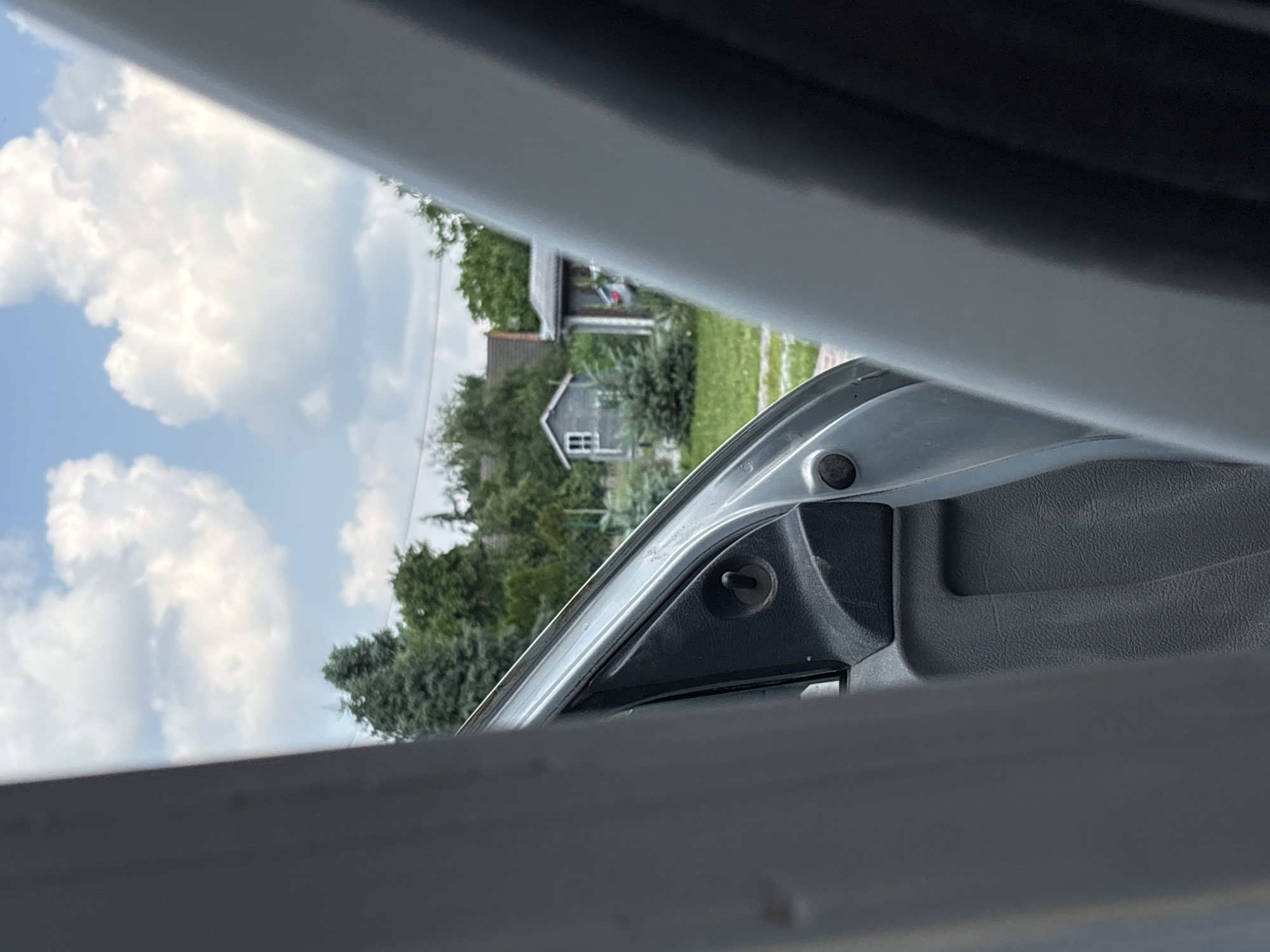 The image depicts a view from inside a vehicle, looking outwards through an open door. The foreground is dominated by the interior of the car door, with the grey plastic panel and a black latch mechanism visible. Beyond the car, a suburban or rural landscape unfolds under a bright, partly cloudy sky. Several trees are scattered across the scene, with two small buildings visible in the background. One building appears to be a small, grey shed with a white-framed window, and the other is a larger, darker structure. The location is identified as Drożki, Poland. The overall impression is that of a snapshot taken while entering or exiting a vehicle in a residential area.