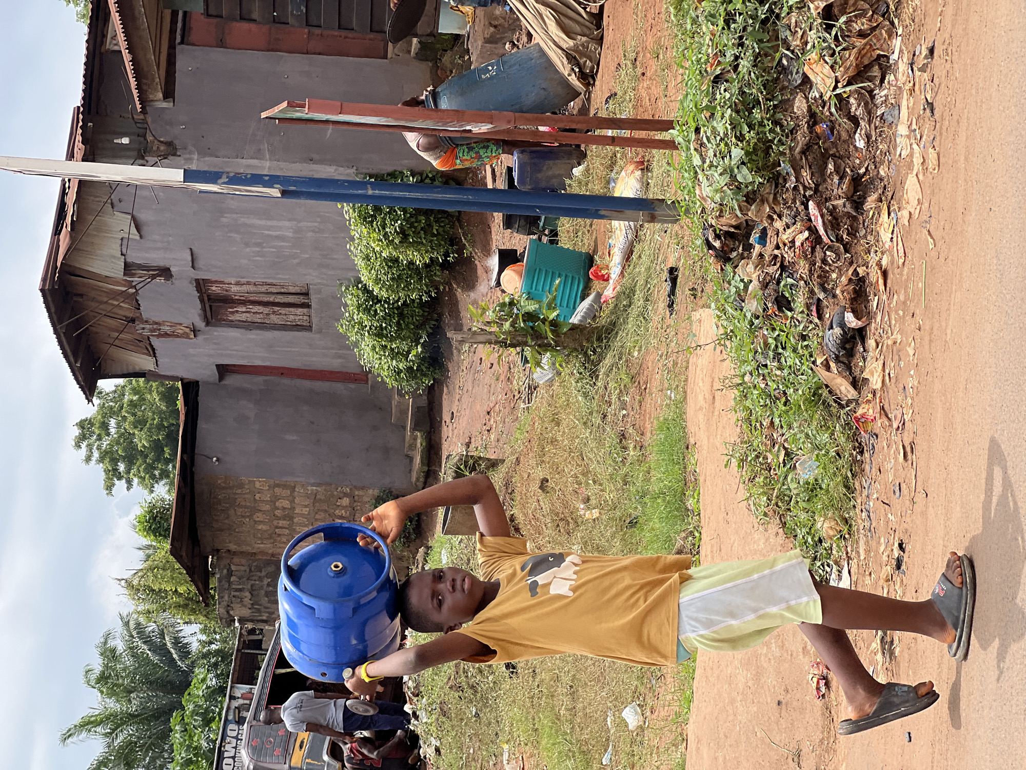 In Nkpor, Nigeria, a young boy with dark skin and short black hair walks along a dusty road, carrying a blue gas cylinder on his head. He is wearing a mustard-yellow t-shirt with a graphic of a black and white cow and light yellow shorts. His feet are clad in dark gray flip-flops. The boy is walking from left to right across the frame, his gaze directed forward.
The environment is characterized by a mixture of residential structures and natural elements. To the left, a yellow truck with "OWO COOKING GAS" written on its side is parked, with several people gathered around it. In the background, buildings with corrugated metal roofs and weathered grey walls are visible. A large pile of green foliage is situated to the right of the boy, with an accumulation of trash and debris at its base. Several blue and red poles stand vertically in the middle of the scene, possibly marking a boundary or pathway. A large blue plastic barrel is positioned behind some of the poles. The ground is a reddish-brown dirt, with scattered patches of dry grass and litter. The sky is overcast with clouds.
