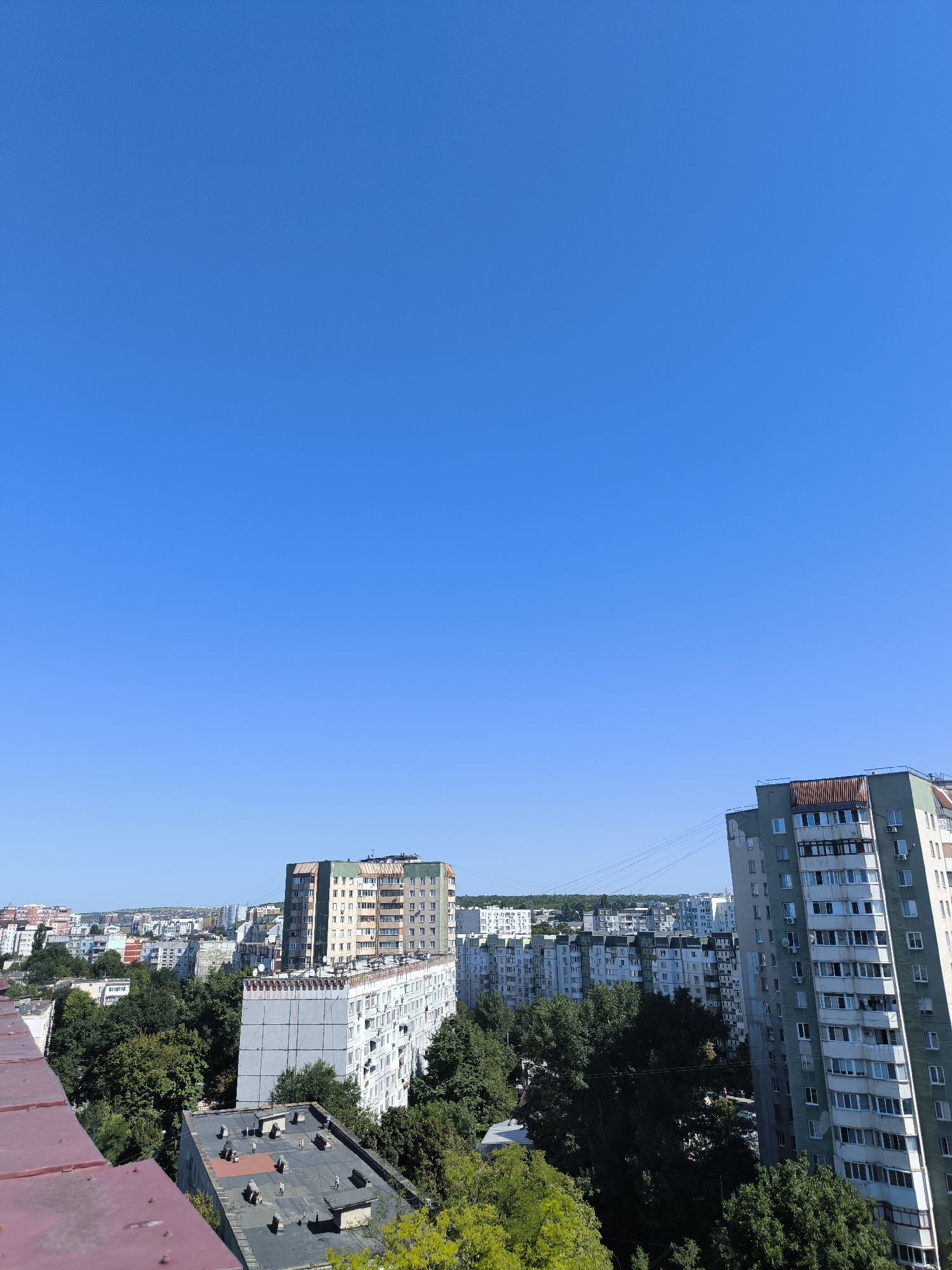 This is a high-angle, medium-shot, eye-level, outdoor photograph taken on a sunny day in Chișinău, Moldova. The scene depicts a dense urban landscape characterized by numerous Soviet-era apartment blocks, predominantly of gray and beige hues. Lush green trees are interspersed throughout the city, breaking up the concrete structures. In the foreground, the edge of a dark red, flat-topped roof is visible, suggesting the vantage point is from a building.
The most prominent structures include a tall, multi-story apartment building on the right, with a light green facade and multiple balconies. Adjacent to it is a wider, lower apartment building with a gray concrete facade and numerous windows. Further into the distance, more apartment buildings of varying heights and colors are visible, creating a layered effect. Some buildings display signs of wear and tear, characteristic of older construction. The sky is a clear, vivid blue with no clouds.
The photograph captures a typical urban panorama of Chișinău, characterized by its post-Soviet architectural style and the presence of abundant greenery. The overall impression is one of a densely populated residential area under a bright, cloudless sky.