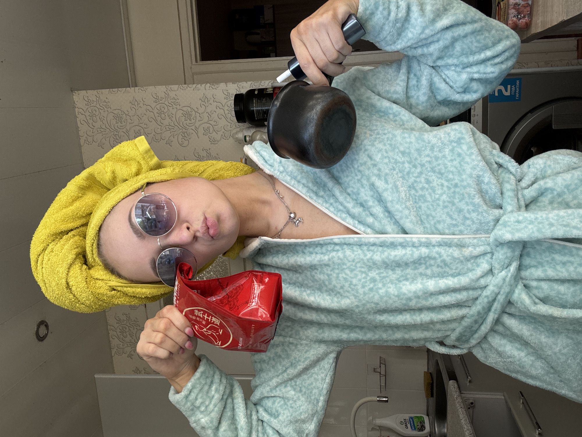A person is wearing a light blue robe, a yellow towel on their head, and round sunglasses. They are holding a black pot with a black spray can attached to it, as well as a red package with Chinese text. The scene appears to be inside a bathroom with white patterned wallpaper, a sink, and a washing machine in the background. Based on the washing machine's signage (гарантии), the image is likely taken in Russia. Further evidence of the location being Chaikovsky is not presented.