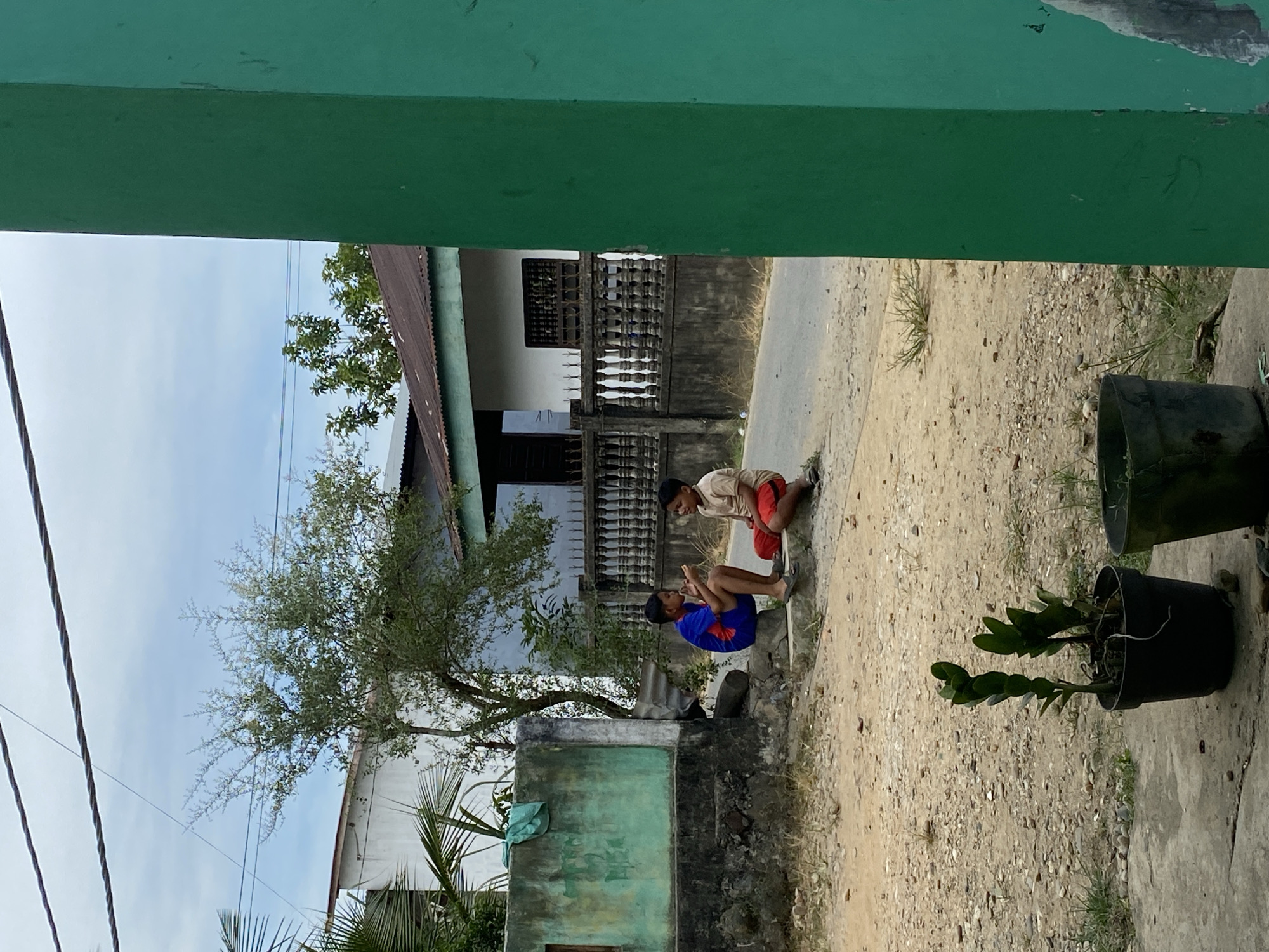 The image displays an outdoor scene, likely in Gampongaceh, Indonesia, framed by a green architectural element, possibly an overhang or balcony, at the top and bottom. Two young boys are central to the image, crouching on a concrete ledge next to a dirt and gravel path. The boy on the left wears a blue shirt and shorts, looking towards the other boy. The second boy, on the right, is in a light-colored shirt and red shorts, also crouching and facing the first boy, suggesting a shared activity or conversation.
In the background, there are houses and buildings, including a white house with a dark, patterned fence and another structure with a weathered greenish-blue wall. A paved road or path is visible behind these buildings. A green leafy tree stands on the left, reaching towards a bright, slightly cloudy sky, with several utility lines visible against it. In the foreground on the right, there are two black potted plants, one containing a leafy green plant. The scene depicts a casual, ordinary moment during what appears to be a clear, sunny day. No visible text is present.