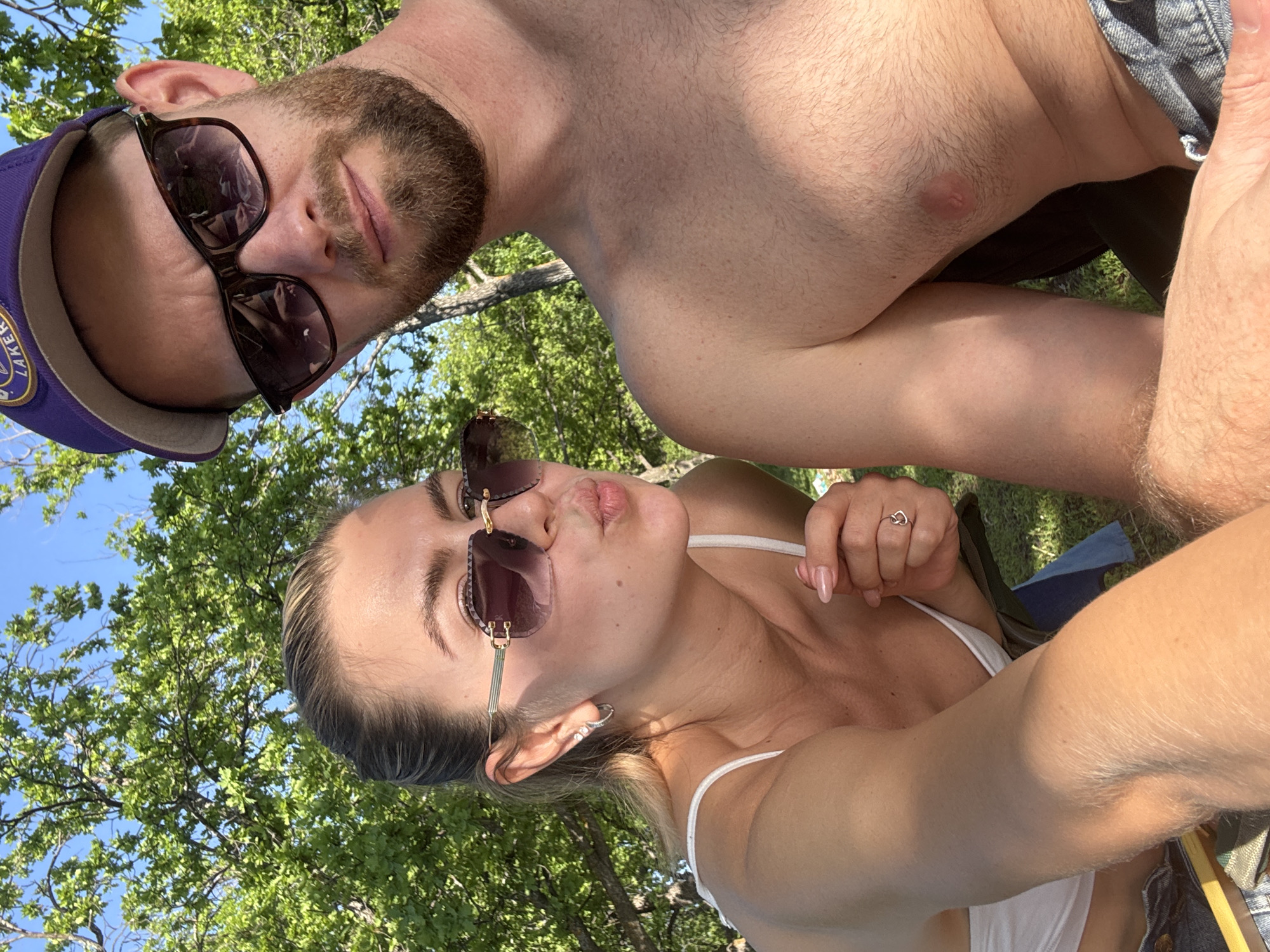 A selfie of a light-skinned man and woman outdoors in Unknown, Unknown. The man has a beard and is wearing a purple Lakers baseball cap and sunglasses. The woman is wearing sunglasses and a white top. There are trees visible in the background.