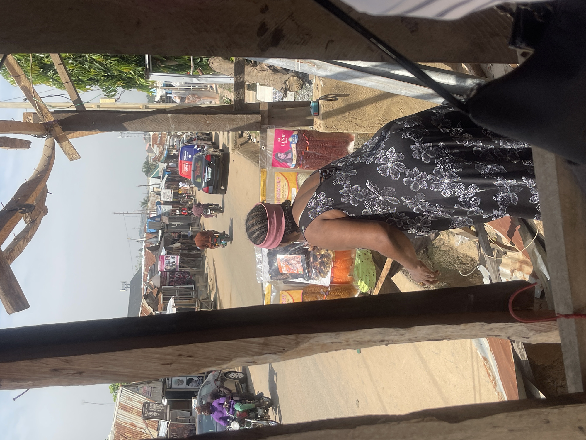 The image shows a street scene in Abuja, Nigeria, likely a market or busy road. The perspective is from under a wooden overhang, looking down and across the street.
In the foreground, a woman is bending over a stall filled with various hair products, including braided extensions in different colors like orange and green, and darker, more natural-looking braids. She wears a black dress with a white floral pattern and a pink headband. Her hand is reaching towards the products.
The street is lined with stalls and small shops. Vehicles, including cars and motorcycles, are visible on the road. One car is red and blue, another is black. A person is riding a motorcycle wearing a purple top. The atmosphere appears to be daytime, with bright sunlight indicating fair weather. The buildings have corrugated metal roofs, common in many Nigerian settings. Signs are visible on some of the shops, although the text is difficult to read clearly. One sign near the hair products appears to advertise "Lush" and "Rosemary."
The overall scene suggests a vibrant commercial area, with people going about their daily activities of shopping and commuting. The image captures a slice of everyday life in a Nigerian city.
