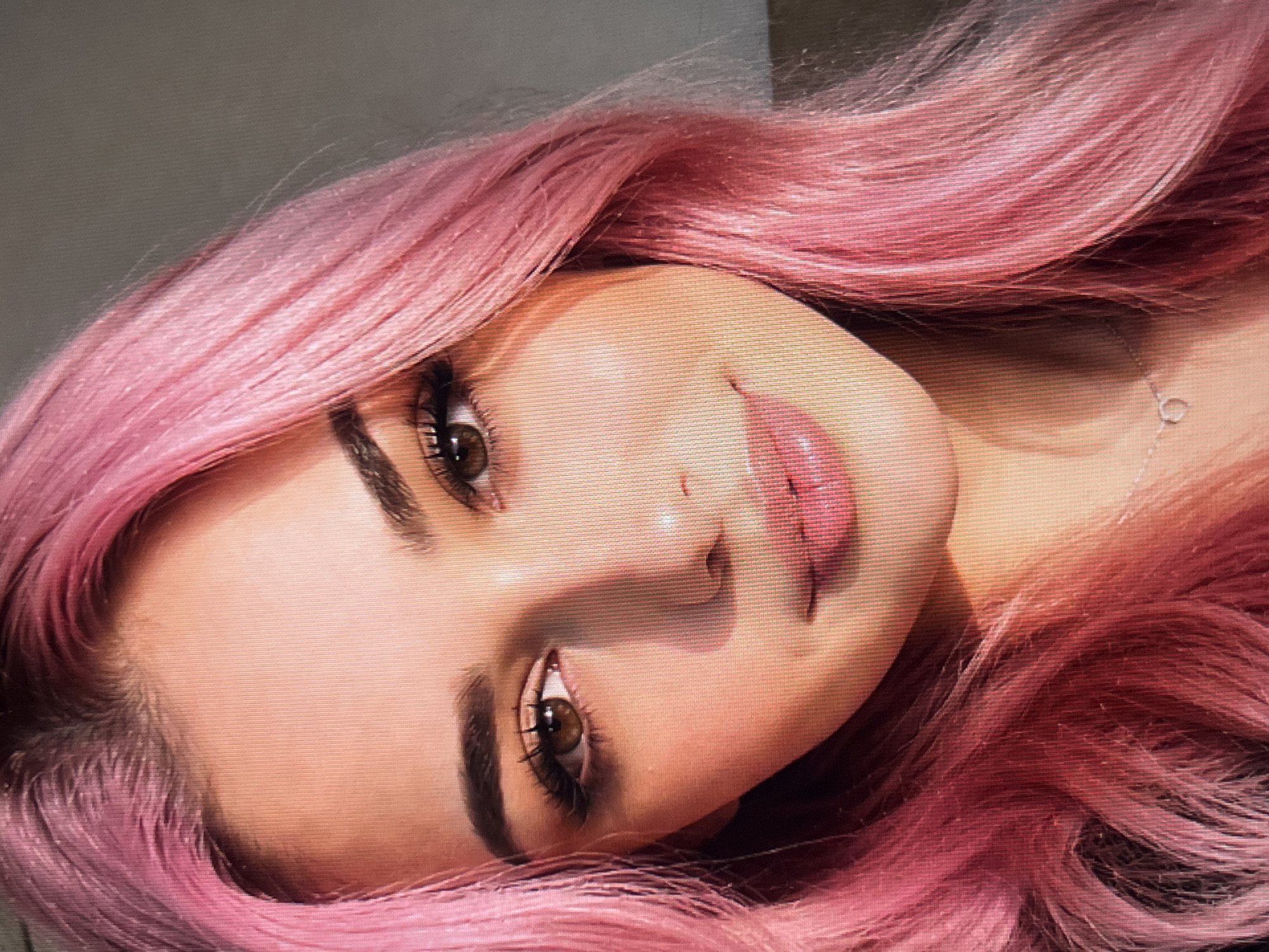 A close-up, headshot photograph shows a young woman with bright pink hair and brown eyes. She is looking at the camera and smiling gently. Her makeup is subtle, with emphasis on her eyes and lips. A thin, silver necklace with a small heart pendant is visible around her neck. The lighting is soft and natural, casting a warm glow on her face. The background is a plain, neutral color. The image is slightly pixelated, suggesting it was taken with a digital camera or smartphone and possibly enlarged.