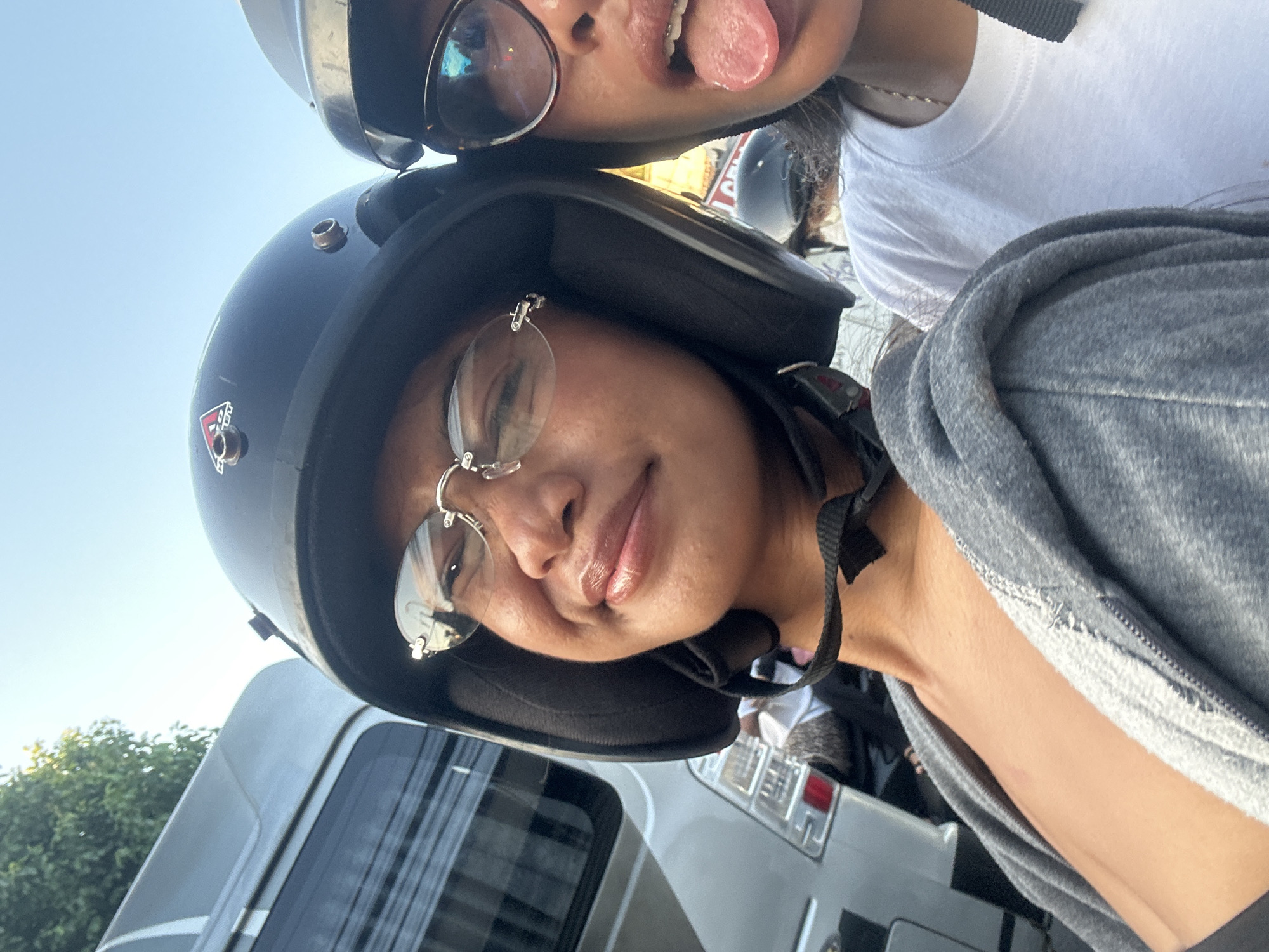 Two women are on a motorcycle in Denpasar, Indonesia. The woman in the foreground is wearing a black helmet and glasses, and she is smiling. The woman behind her is wearing sunglasses and sticking her tongue out. They are both wearing casual clothing. In the background, there is a silver van and some trees. The sky is clear and blue.