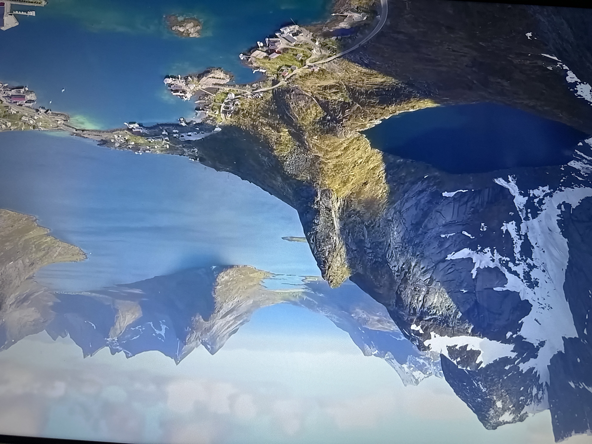 The image displays an upside-down view of a coastal landscape, likely a fjord or series of islands and mountains.
**Environment:** The scene includes a body of water, possibly the ocean or a fjord, with several islands and peninsulas. The land features are characterized by rugged, rocky mountains with some snow-capped peaks. There are areas of green vegetation on the slopes.
**Subjects:** There are several small settlements or villages visible along the coastlines of the islands and peninsulas.
**Objects:** Houses and buildings are present in the settlements. Roads or bridges connect some of the islands. There are also visible piers/docks for boats.
**Actions:** The scene captures a static view of the landscape with no visible actions or interactions.
There is no indication that the picture includes information about the city of Bragança, Portugal.