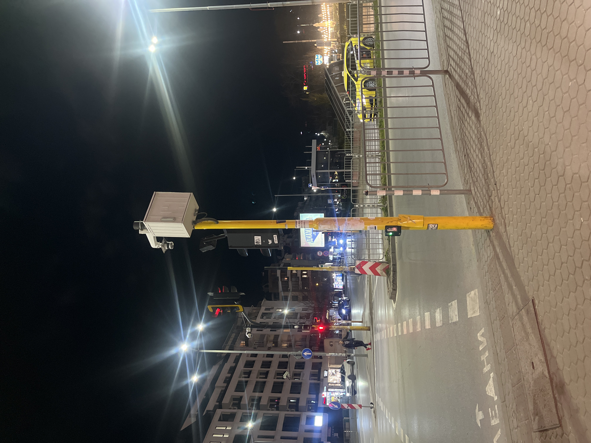 The image shows a street scene at night in Sofia, Bulgaria.
Foreground: A yellow traffic light pole is prominently featured on the right. A pedestrian walkway with hexagonal paving stones is visible.
Middle Ground: A street with lane markings leads away from the camera. A pedestrian is crossing the street. Cars are driving through the intersection. Traffic lights are visible, some displaying red and some green. A multi-story building is present.
Background: Street lights illuminate the scene. In the distance, signage and building lights create a blurred cityscape. The Cyrillic alphabet is visible on some signs.
Overall: The image captures a typical urban night scene with pedestrians, vehicles, and traffic infrastructure. The city of Sofia is the location.