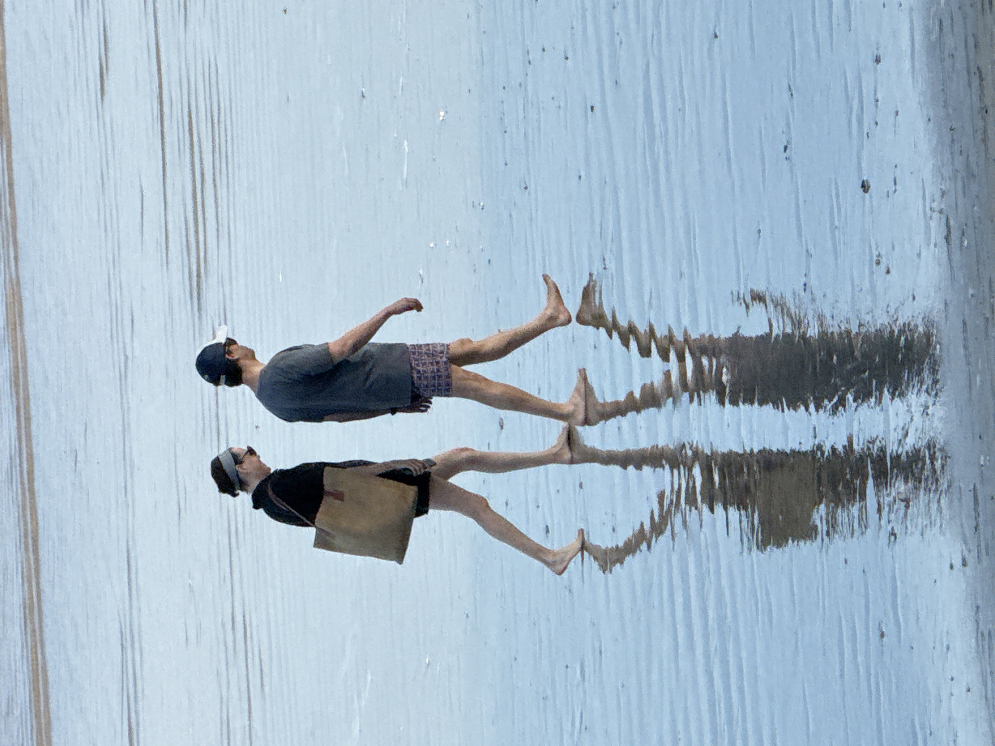 Two individuals are walking side-by-side on a wet, sandy beach in Kuta Selatan, Indonesia. The person on the left, appearing to be female, wears a dark hooded jacket, a headband, and sunglasses. They carry a large, light-brown tote bag over their left shoulder. The person on the right, appearing to be male, wears a dark t-shirt and patterned shorts, with a dark baseball cap and sunglasses. Both are barefoot and their reflections are clearly visible on the wet sand. The water is shallow and calm, with faint ripples creating a textured surface. The background shows the expanse of the beach and possibly the ocean or a lagoon.