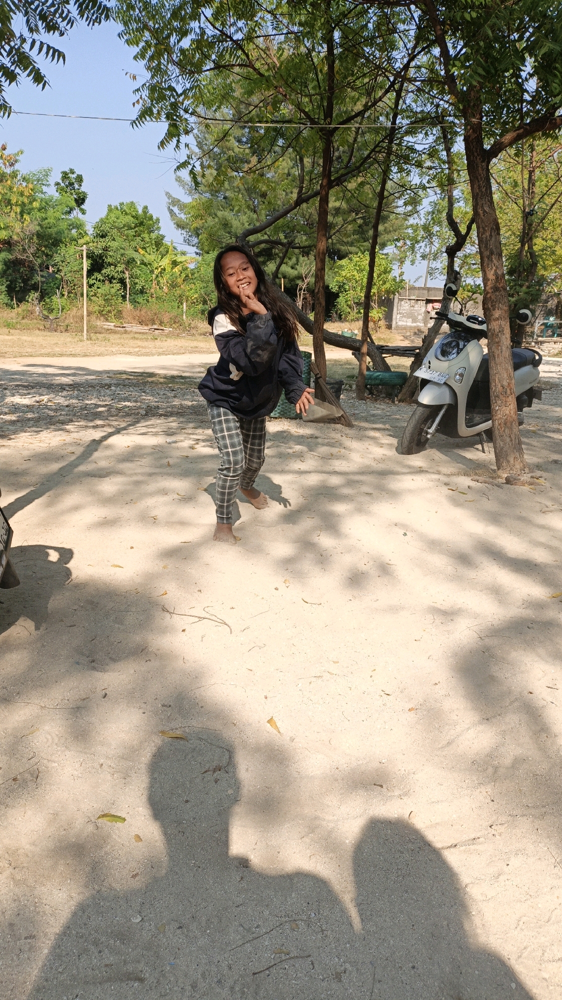 A young girl is standing outdoors in the city of Cikal, Indonesia, on a sandy path shaded by trees. She is wearing a dark blue jacket with white accents and grey checkered pants. Her right hand is raised to her mouth, and she is smiling and looking at the camera. A silver scooter is parked behind her. The ground is covered in sand with fallen leaves and twigs. Shadows of trees and people are cast across the ground. The sky is clear and blue.