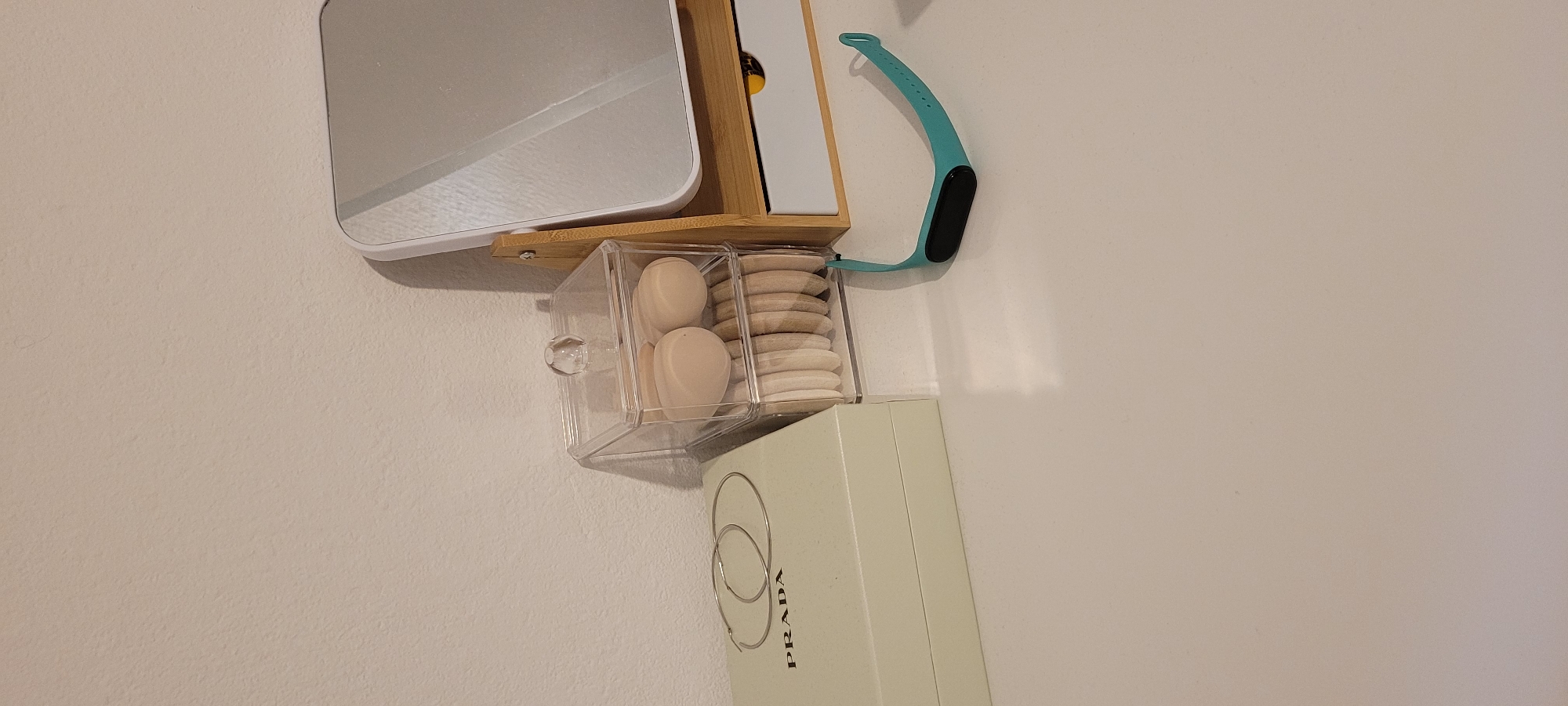 The image depicts a static, eye-level view of a well-organized personal grooming or vanity area against a light beige textured wall and atop a white, smooth, reflective surface. The scene offers no discernible visual information to confirm a location in the city of Heusenstamm, Germany. No individuals are present.
On the left, a light mint green rectangular box, marked with "PRADA" in black capital letters on its front face, is positioned. Two thin, silver-colored hoop earrings rest diagonally on its lid.
Adjacent to the Prada box, a clear, two-tiered acrylic organizer holds various makeup applicators. The upper compartment contains two beige, egg-shaped makeup sponges. The lower compartment is filled with multiple light beige, circular, flat powder puffs or sponges, neatly stacked vertically. A small, clear, spherical knob serves as a handle on the organizer's lid.
To the right of the organizer, a white-framed rectangular mirror with rounded corners stands upright on a light wooden (possibly bamboo) stand. The stand features a small white drawer at its base, from behind which a small, bright yellow object is partially visible. The mirror reflects a bright light source, indicating an illuminated indoor environment.
In the foreground, on the white surface to the right of the Prada box and in front of the mirror stand, lies a teal-colored fitness tracker band with its black rectangular sensor/display unit inserted. The band is curved, suggesting it was recently removed or placed.
