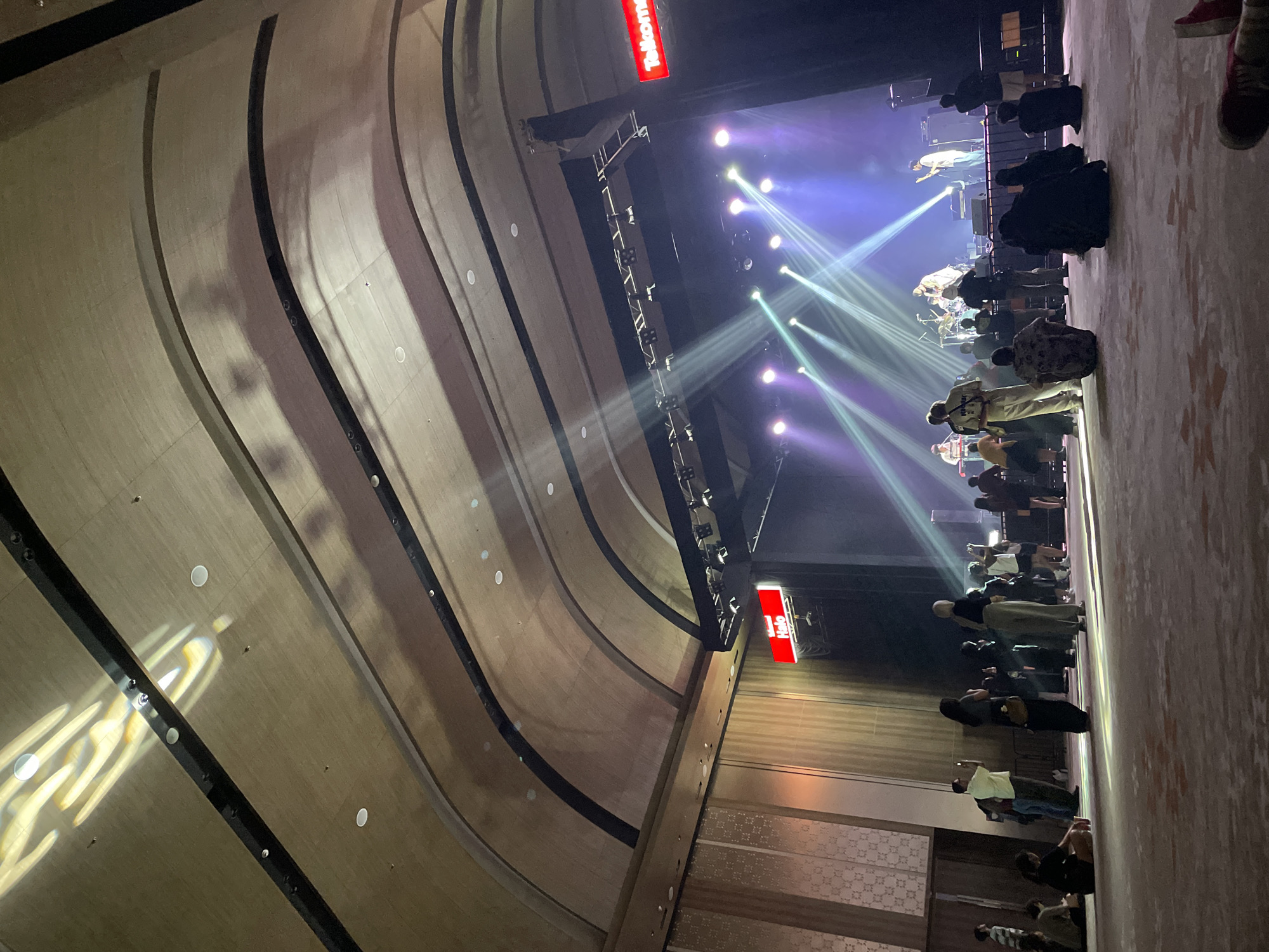 Here is a description of the image:
The image shows a concert hall in Salembaran, Indonesia, likely during a performance. The perspective is from a low angle, looking towards the stage. The hall has a distinctive curved ceiling with repeating linear patterns and embedded lights. On stage, musicians are performing under bright spotlights creating beams of light that cut through the air. A crowd of people is gathered in front of the stage, watching the performance. The Telkomsel logo is visible on a sign.