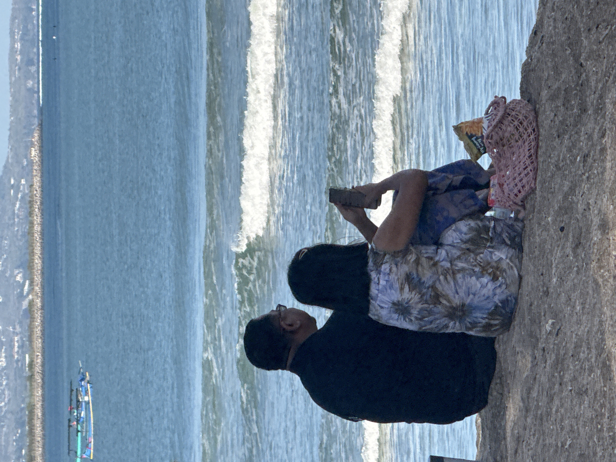Two individuals, a man and a woman, are seated on a sandy incline overlooking the ocean in Kuta, Indonesia. The man, wearing a black collared shirt, is positioned to the left, his back to the viewer. He appears to be wearing glasses. The woman, seated to his right and slightly in front, is wearing a patterned blue and white top and blue patterned pants. She is holding a smartphone in her right hand, seemingly looking at it. In front of her on the sand is a pink mesh bag containing a bag of chips. The ocean stretches out before them, with gentle waves rolling towards the shore. In the distance, several boats are visible on the water, and a coastline with buildings and vegetation is discernible on the horizon under a clear sky.