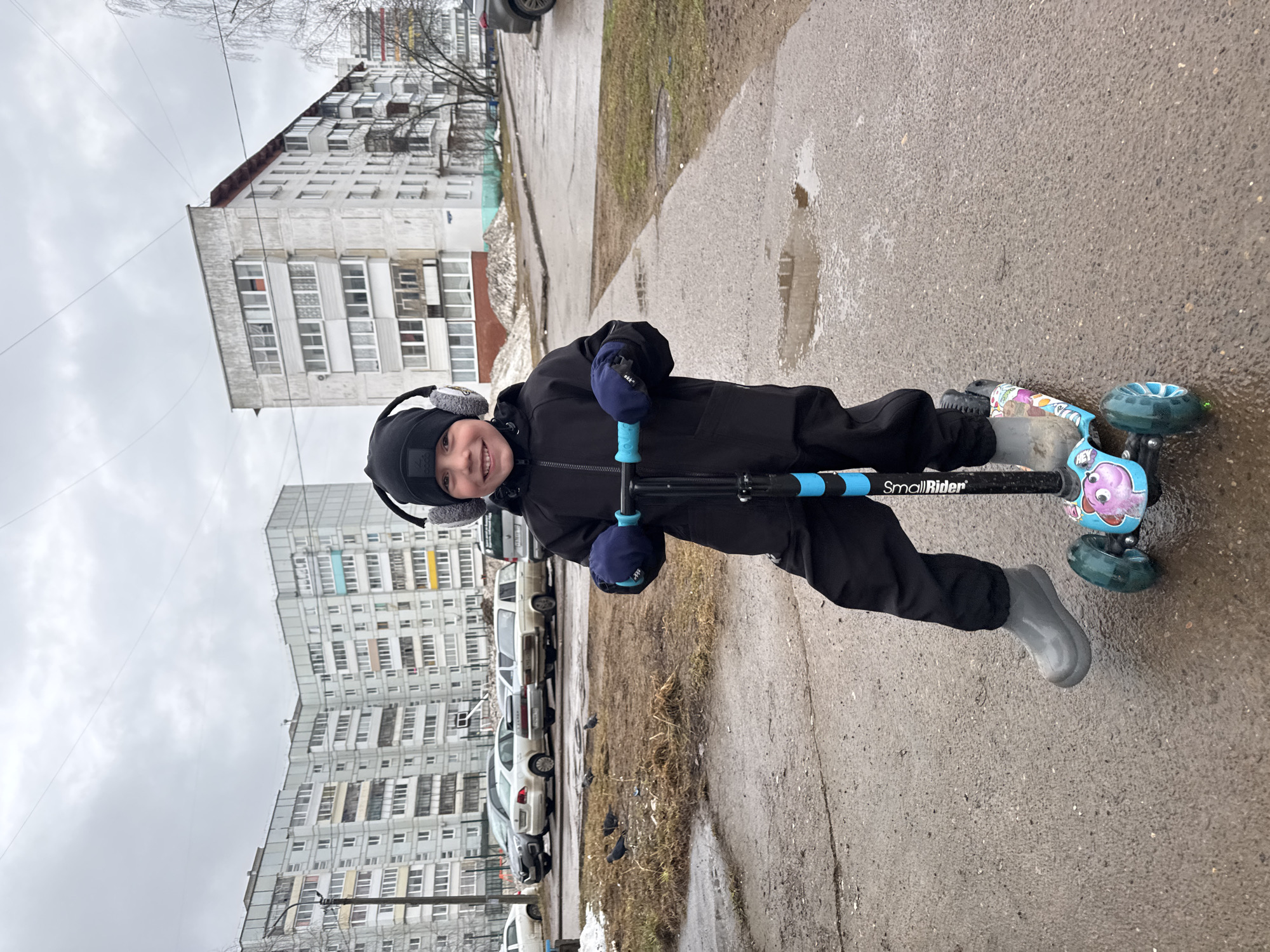 A child on a scooter is positioned on a paved surface, likely a sidewalk or street, in what appears to be the city of Chaikovsky, Russia, based on the urban architecture. The child is wearing a black jacket, pants, hat with earmuffs, and gloves, with gray-colored boots. The scooter is black with blue handles and features the words "Small Rider." The background shows a large multi-story apartment building and parked cars. The weather appears overcast and damp, with visible puddles on the pavement.