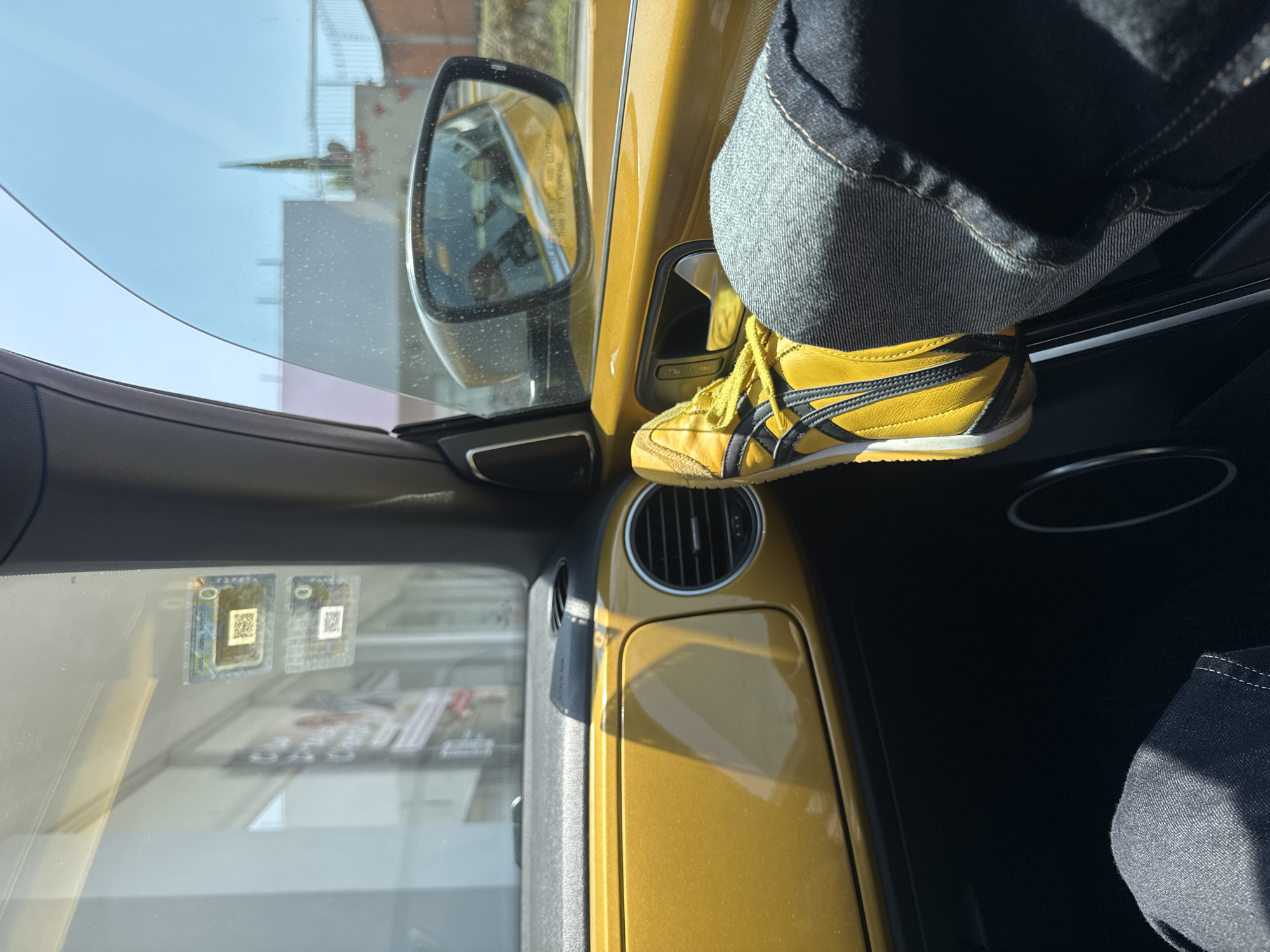 A shoe on the door of a yellow vehicle. The shoe is yellow with black stripes and yellow laces. A leg in blue jeans is visible next to the shoe. Through the vehicle's windows, a streetscape is visible. Based on the stickers on the windshield, the location is Puebla, Mexico.