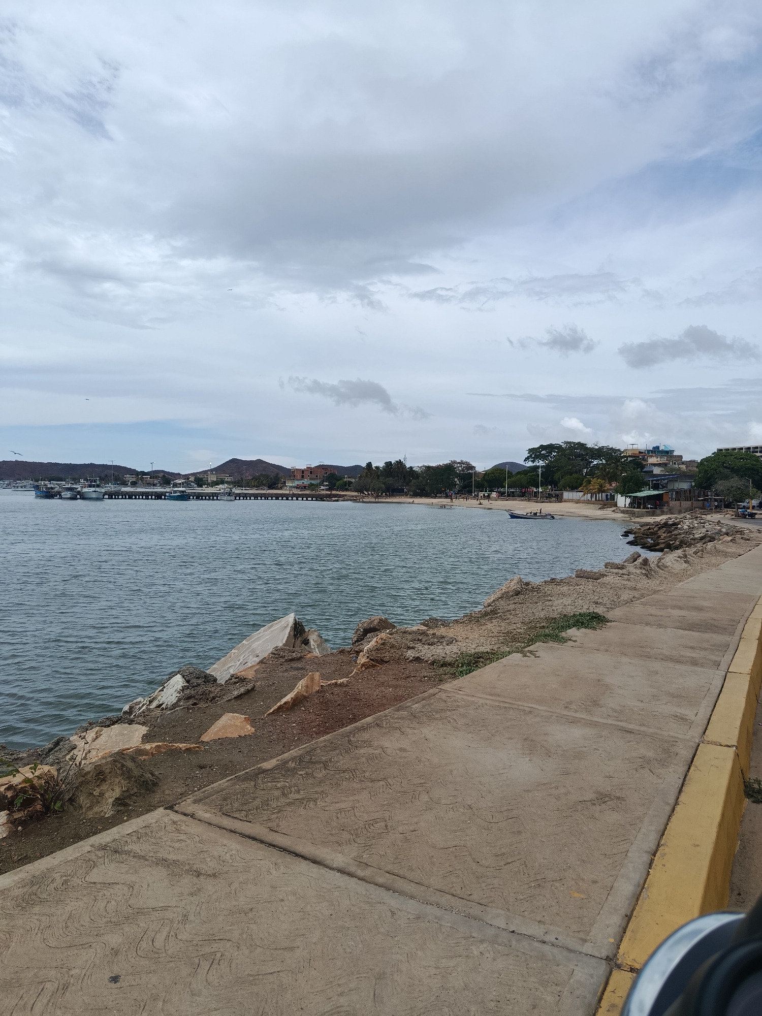 This image depicts a coastal scene in Juan Griego, Venezuela, under an overcast sky. In the foreground, a textured concrete pedestrian walkway with a distinctive wave pattern runs from the lower left to the right, bordered by a yellow-painted curb on its right edge. To the left of the walkway, a rocky, dirt embankment slopes down to the water, displaying sparse vegetation.
The middle ground features a calm, dark blue-grey bay with gentle ripples. On the left side of the bay, a long pier or jetty extends, alongside which several small, light-colored boats are moored. Beyond the pier, low, arid hills are visible, dotted with some buildings. Towards the center and right, a sandy beach curves, where one small blue and white boat is pulled onto the sand. Scattered individuals can be seen on the beach. Behind the beach, mature trees line the shore, and various single and multi-story buildings with colorful exteriors are present, indicating a small coastal settlement. Rocky formations delineate the transition from the paved area to the beach on the right.
The background is dominated by a heavily overcast sky, filled with uniform light grey clouds. Distant, low, dry hills complete the horizon. A small, dark speck, possibly a bird, is visible in the upper central sky. The overall lighting is diffused, consistent with a cloudy day.