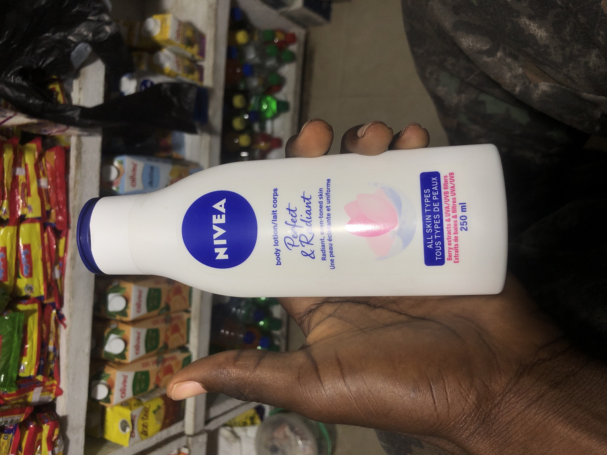 A person holds a white NIVEA Perfect & Radiant body lotion bottle in front of a shelf stocked with various products. The bottle, with a blue cap and label, features text in English and French. The background shows shelves filled with packaged goods, including what appear to be juice boxes and snacks. The setting suggests a retail environment, possibly a small shop or market stall in Ado, Nigeria, given the presence of common consumer products. The lighting is dim, typical of an indoor setting with limited natural light. The person's hand is dark-skinned and is positioned to cradle the bottle. The overall scene is a candid snapshot of a product being held up for display or inspection.