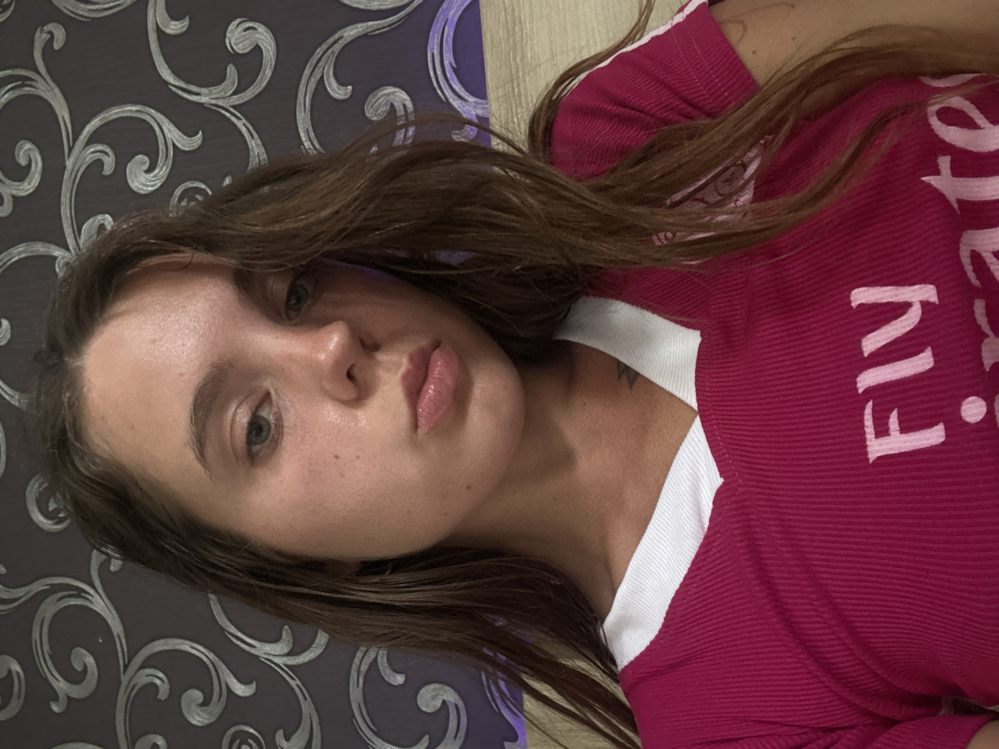 An indoor, close-up, selfie-style, medium shot captures a young Caucasian woman from the chest up. She has long, wavy brown hair that falls around her face and shoulders. Her skin appears slightly flushed and dewy. She is wearing a bright pink, ribbed t-shirt with a white V-neck collar. The t-shirt has white lettering on it, with the partial word "Fly" visible. She is looking directly at the camera with her lips pursed. The background is a dark grey wallpaper with a swirling, ornate silver pattern. A hint of a light-colored surface, possibly a bed or furniture, is visible at the bottom of the frame. The lighting is soft, illuminating her face and upper body.
