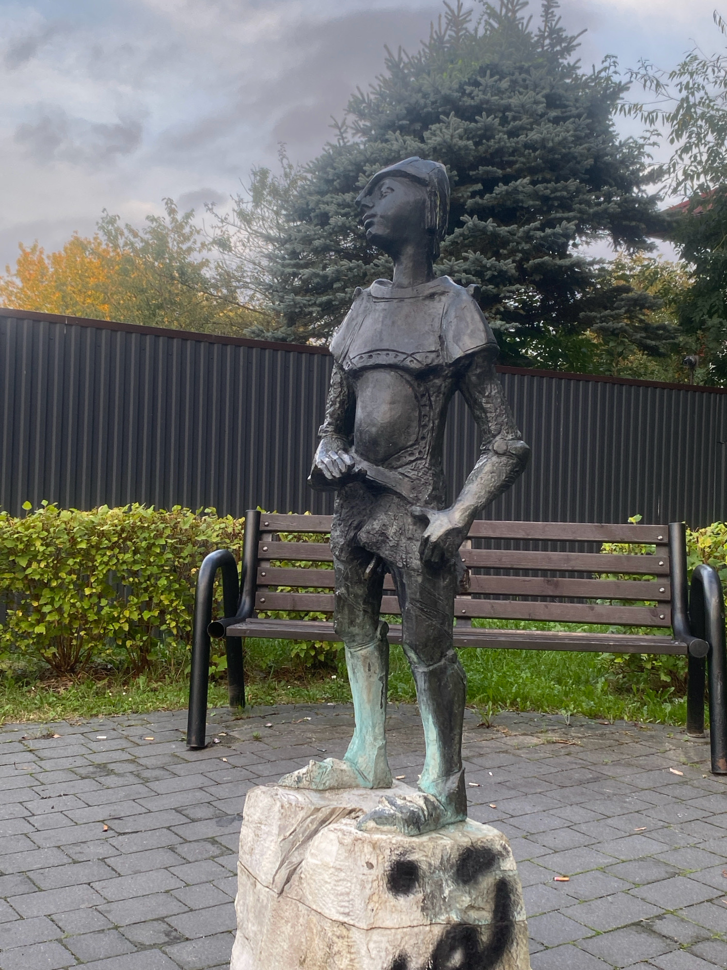 This image depicts a bronze statue of a young boy dressed in armor, standing on a stone pedestal. The statue is located in a park-like setting in Nowy Sącz, Poland. The boy is depicted in a dynamic pose, looking to his left with his mouth slightly open. He is wearing a helmet, chest plate, arm guards, and greaves. His right hand is holding a sword, and his left hand rests on his hip.
Behind the statue, there is a wooden bench with black metal legs, a lush green hedge, and a dark gray corrugated metal fence. A large fir tree is visible in the background, along with other foliage and a cloudy sky. The ground in front of the statue is paved with gray rectangular bricks. The stone pedestal appears to have graffiti on its front surface. The overall impression is that of an outdoor public space, possibly a memorial or a decorative element in a park or square.