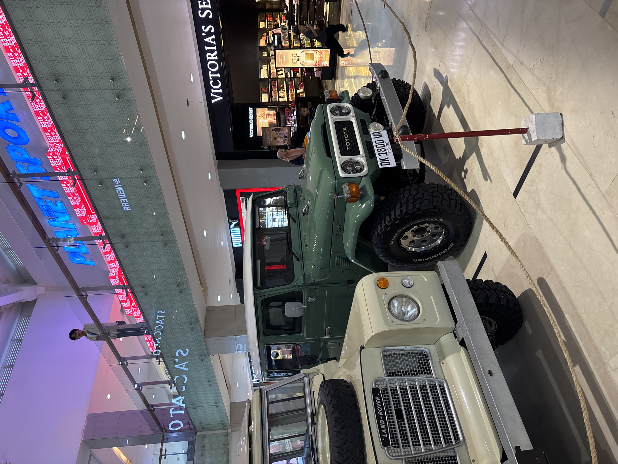 Here's a concise description of the image:
The image shows two vintage off-road vehicles, a green Toyota Land Cruiser and a beige Land Rover, displayed inside a shopping mall. The vehicles are positioned next to each other and are roped off for display. The Toyota has a license plate reading "DK 1800 VA". Visible in the background are storefronts for "VICTORIA'S SECRET" and other shops. The mall has multi-level architecture, with people seen on the upper levels. Store signage like "PLANET SPORTS" is visible. The picture was taken in the city of Kuta, Indonesia.