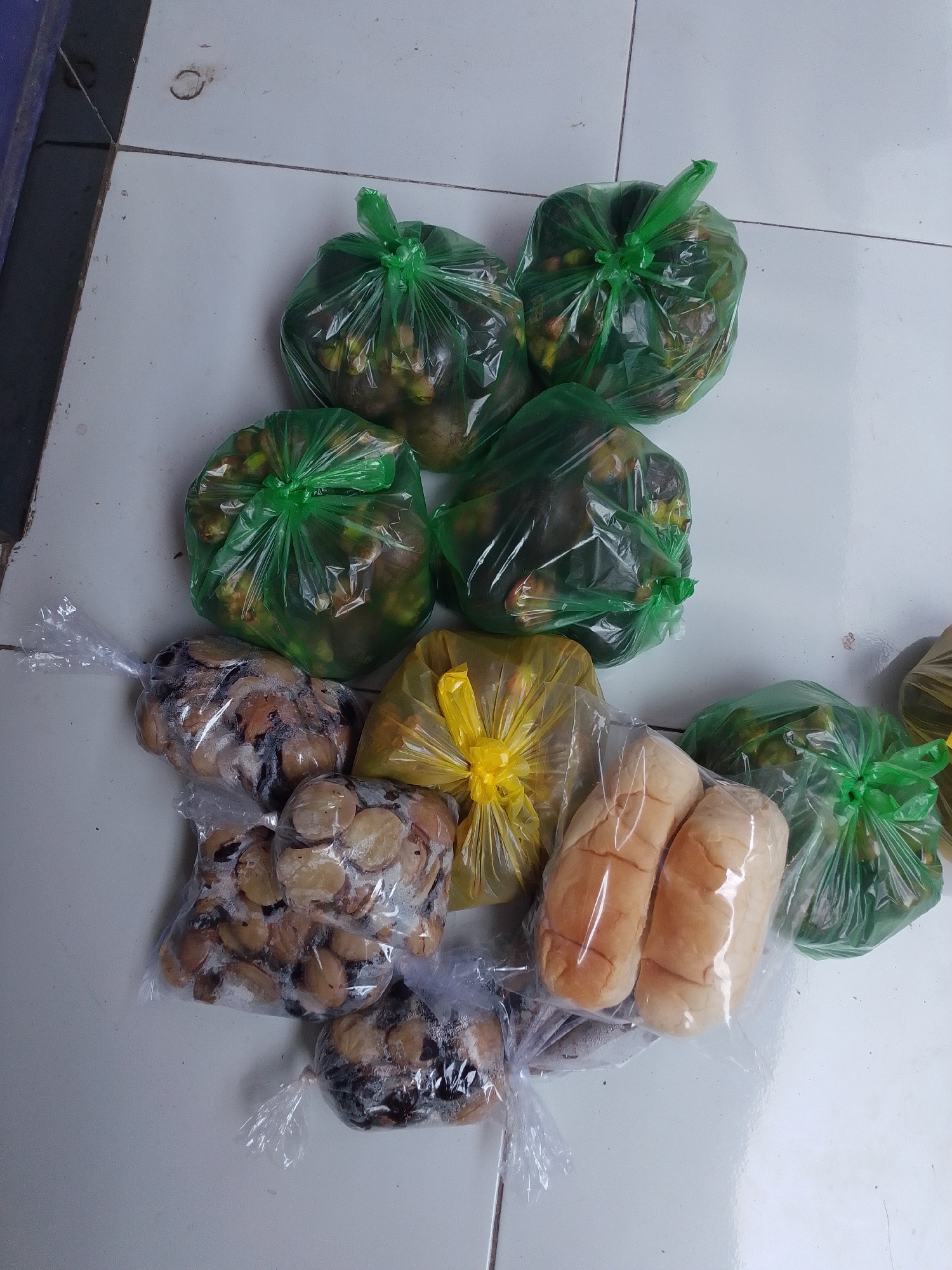 In Panyingkiran, Indonesia, on a tiled floor, several plastic bags filled with food items are arranged. Three green plastic bags contain what appear to be fruits or vegetables, possibly mangosteens, given their rounded shape and the presence of stems. To the left, two clear plastic bags hold numerous sliced pieces of food that resemble dried tamarind. A yellow plastic bag with a tie closure contains more of the green bagged items. In the foreground, two loaves of bread are visible within a clear plastic bag, and another clear plastic bag below contains more of the sliced, dried food. The scene suggests a collection of groceries or prepared food items.