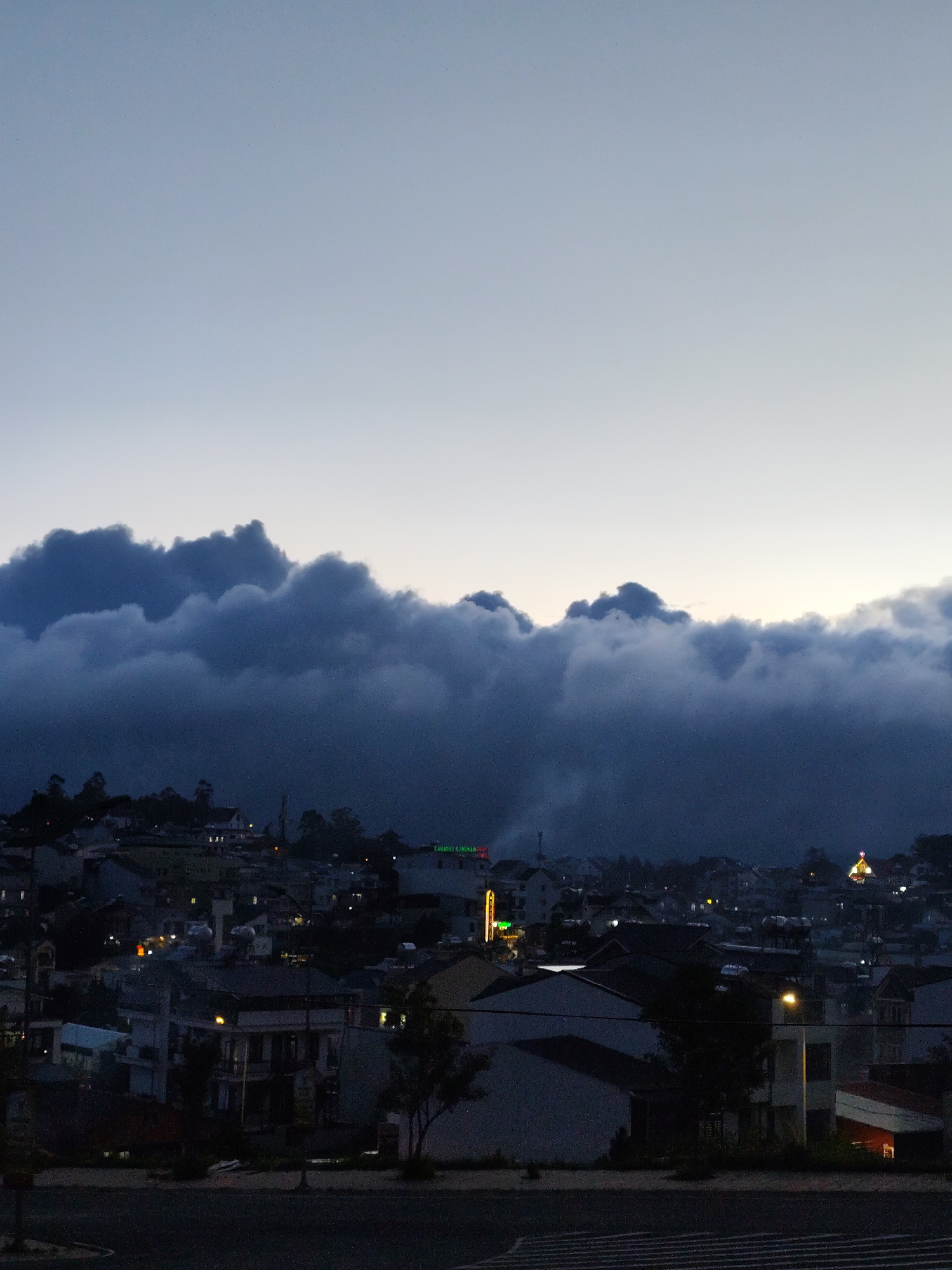 This is a nighttime scene of a city in Lang Biang - Da Lat, Vietnam. The sky transitions from a light blue to a darker hue with thick clouds in the midground. Below the clouds, the city unfolds with buildings illuminated by various lights, including neon signs. The foreground is dark, with a road surface showing striped lines. The overall atmosphere suggests a cool, possibly foggy evening.