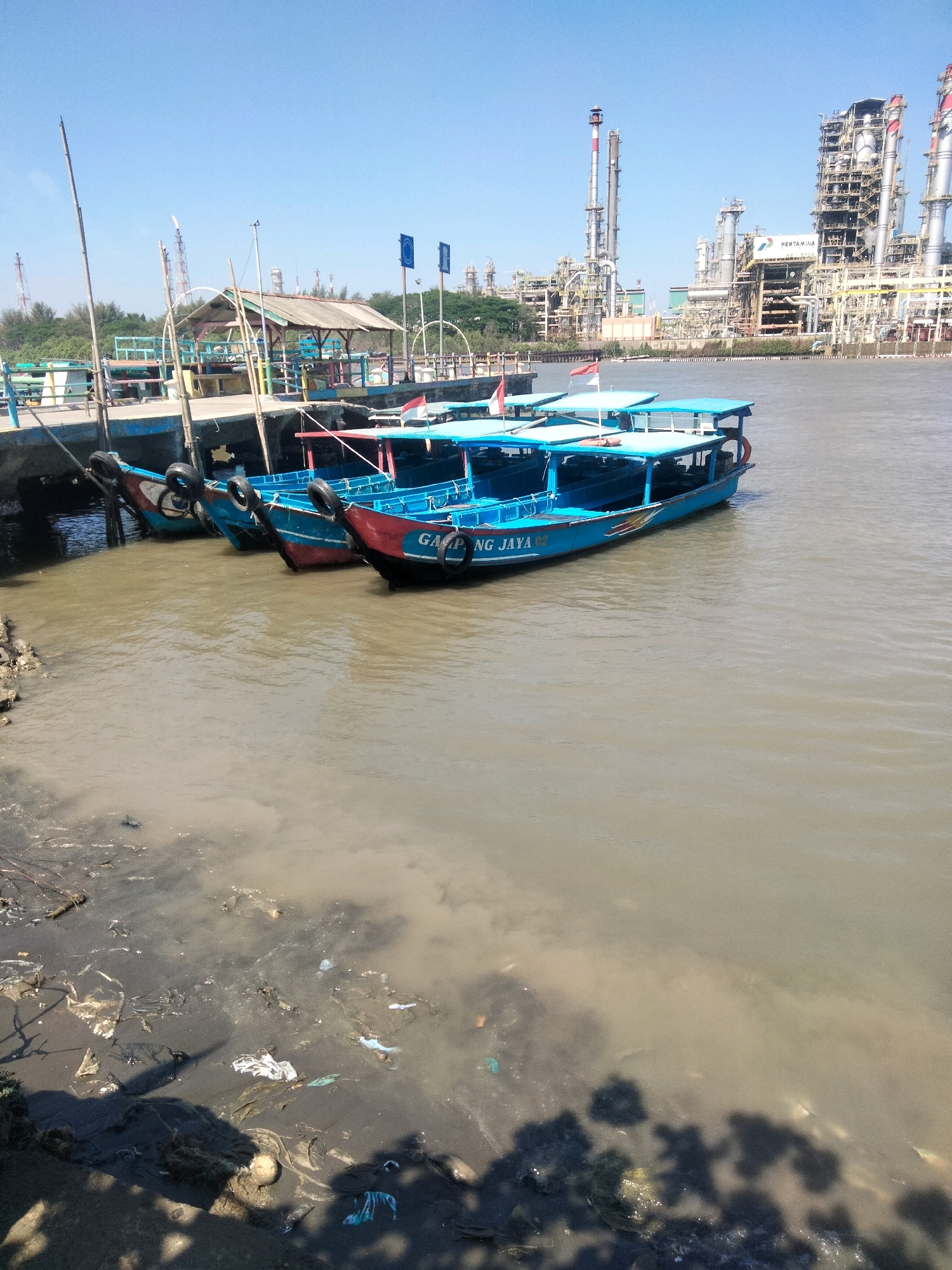 The image depicts a waterfront scene in Cilacap, Indonesia, likely near the Pertamina refinery as suggested by the industrial structures in the background. In the foreground, several blue and red passenger boats are docked at a pier. The water is murky and polluted, with visible trash along the shoreline. The sky is clear and blue. The overall impression is one of a working port area with visible environmental concerns.