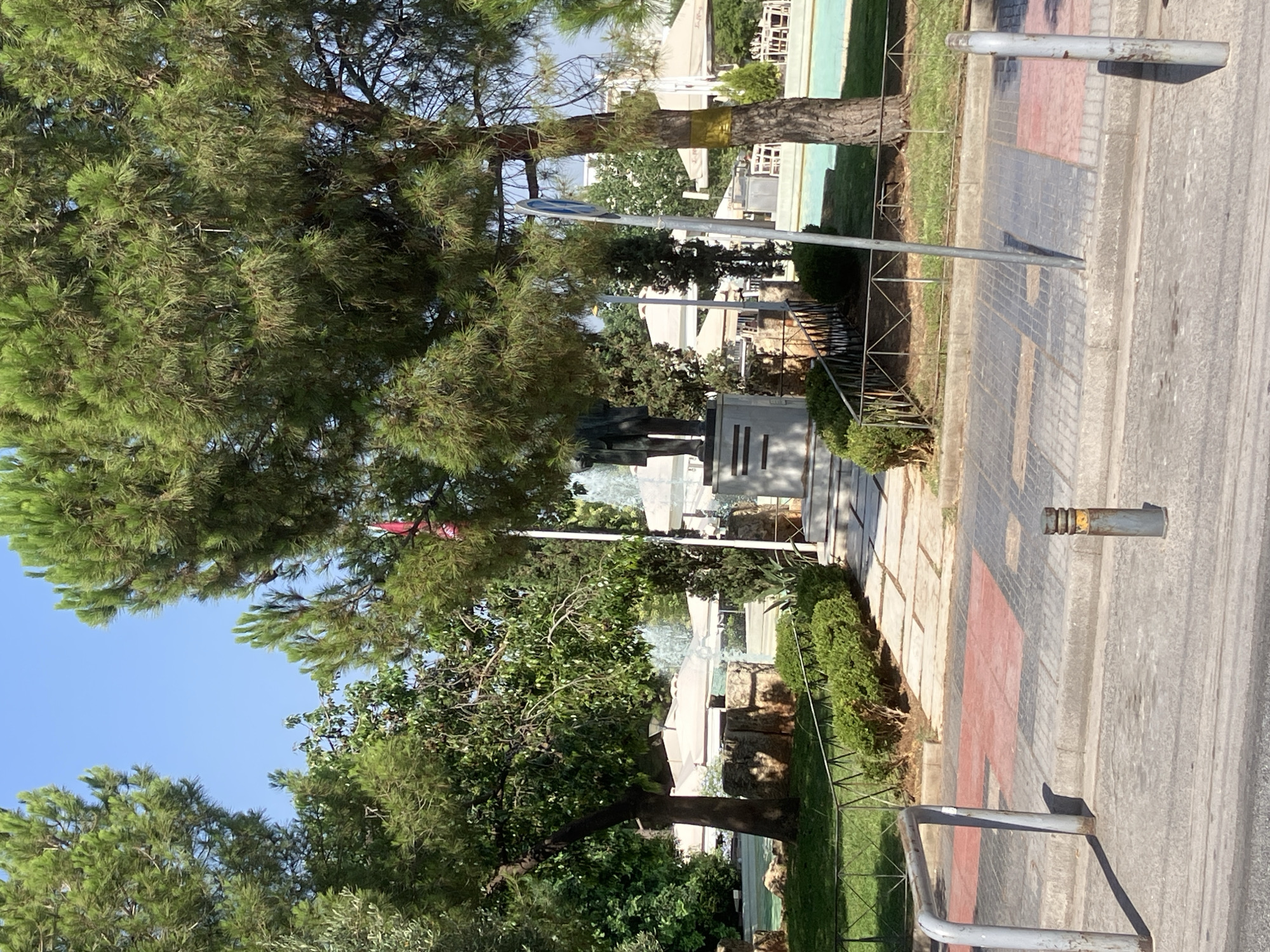 This image captures a sunlit scene in Nea Smirni, Greece, focusing on a public square. In the foreground, a gray concrete sidewalk with red and gray paving stones lines the street. Two sturdy metal bollards are positioned on the sidewalk, one to the left and one to the right, appearing to serve as barriers or decorative elements. A gray metal railing separates the sidewalk from a grassy area.
Beyond the railing, a paved pathway leads towards a monument. The monument features a statue of a man in a suit, standing atop a stone pedestal. Behind the monument, lush green trees provide shade, and in the background, white umbrellas suggest outdoor seating areas, possibly for a cafe or restaurant. A bright blue body of water, possibly a pool or fountain, is visible in the mid-ground. The sky is a clear, bright blue. The overall impression is one of a well-maintained public space on a sunny day.