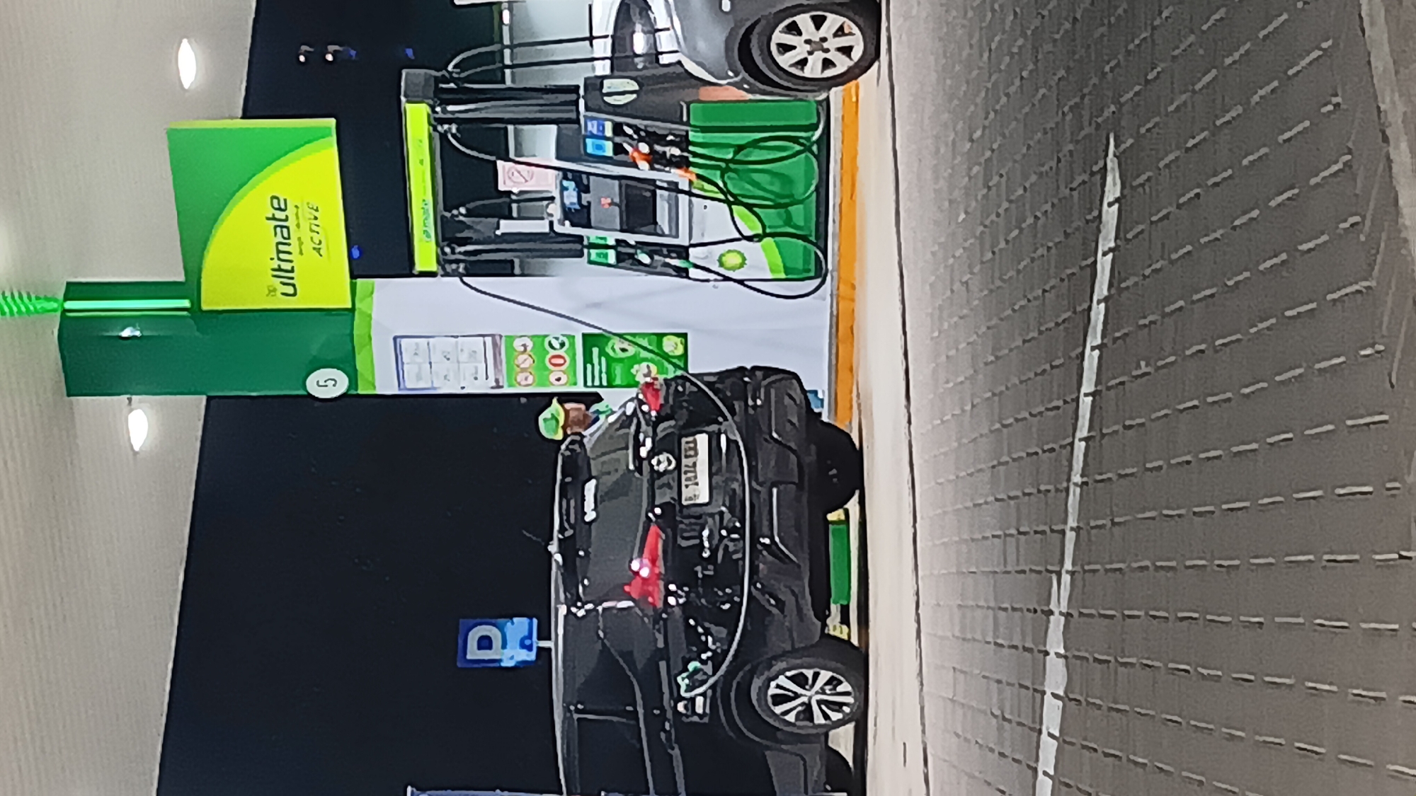 An outdoor night scene at a BP gas station in South Jakarta, Indonesia shows a black SUV being refueled. The SUV has a license plate that reads "B 1874 EXX". A person wearing a green cap and green shirt is standing near the gas pump. The gas station has BP branding, including a prominent "ultimate ACTIVE" sign. The ground in the foreground is a patterned paved surface, and the background is dark, suggesting it is nighttime. Another car, partially visible on the right, is also at the gas station.