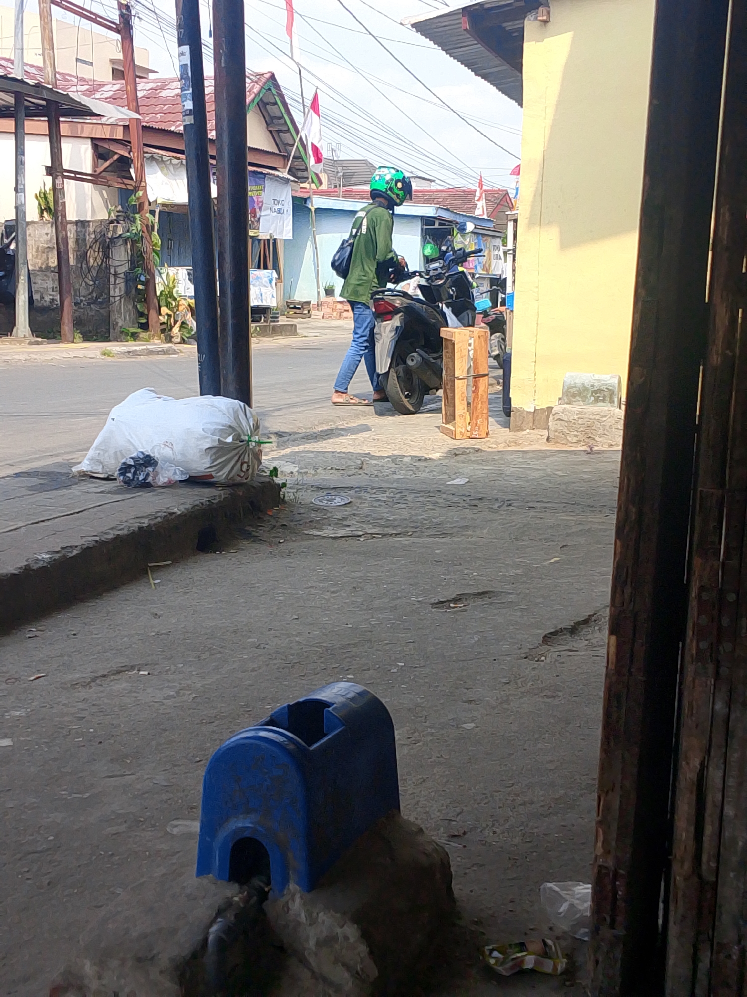 The image shows a street scene in Palembang, Indonesia. In the foreground, there is a blue plastic trash can on the sidewalk, with trash overflowing from it. To the left, a large white garbage bag is placed on the curb. In the middle ground, a person wearing a green jacket and a green helmet is standing next to a motorcycle. The motorcycle has a wooden crate on its side. Behind the person and motorcycle, there are several buildings with red tile roofs. A red and white flag is flying from one of the buildings. In the background, there are utility poles and power lines. The ground is paved with asphalt and concrete. The lighting suggests it is daytime.