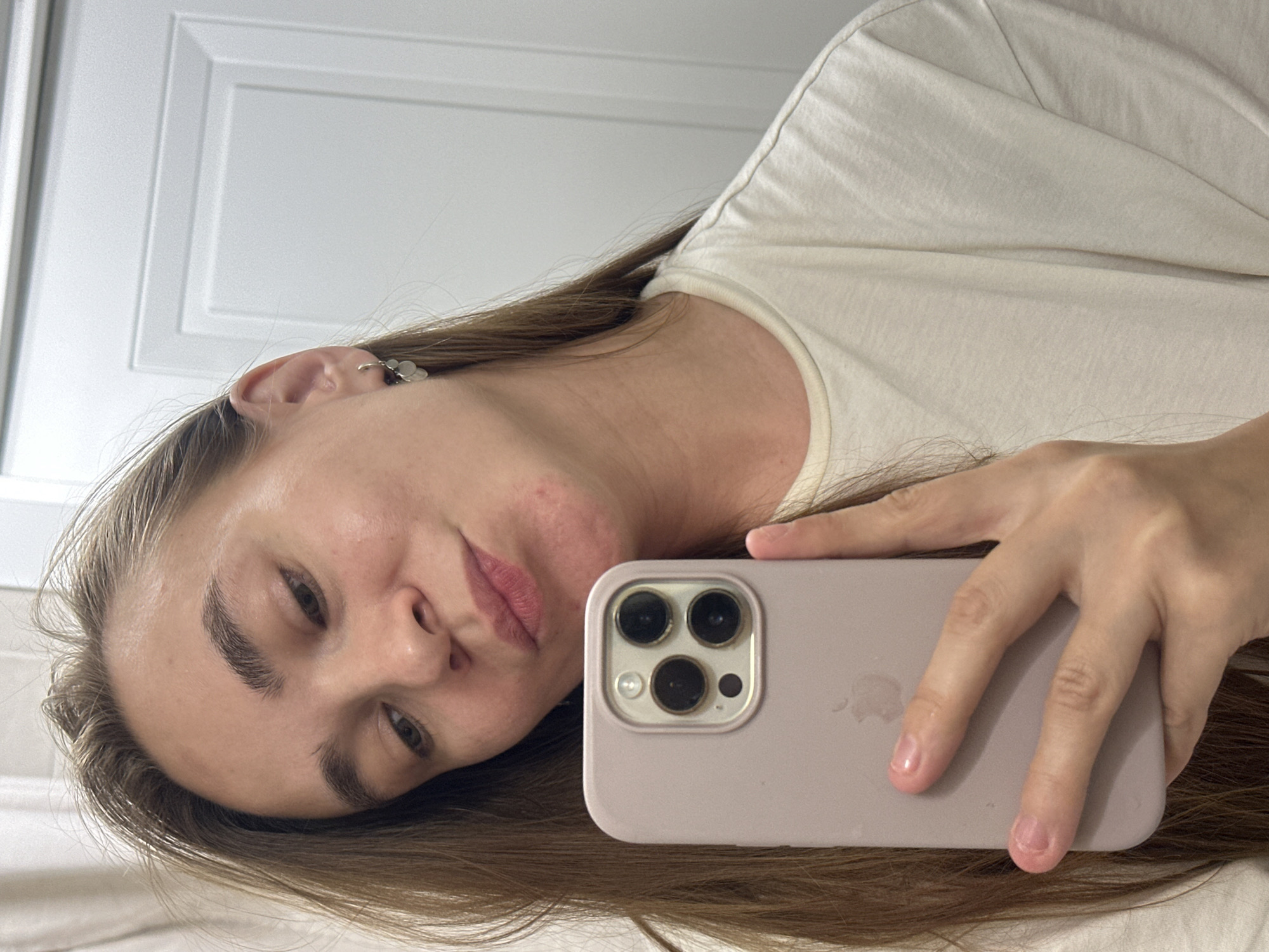 A woman is shown in a selfie, holding a phone with three camera lenses and a light lavender case. She wears a light-colored shirt and has light brown hair. An earring is visible on her left ear. The background includes a white paneled door. The location is not specified.