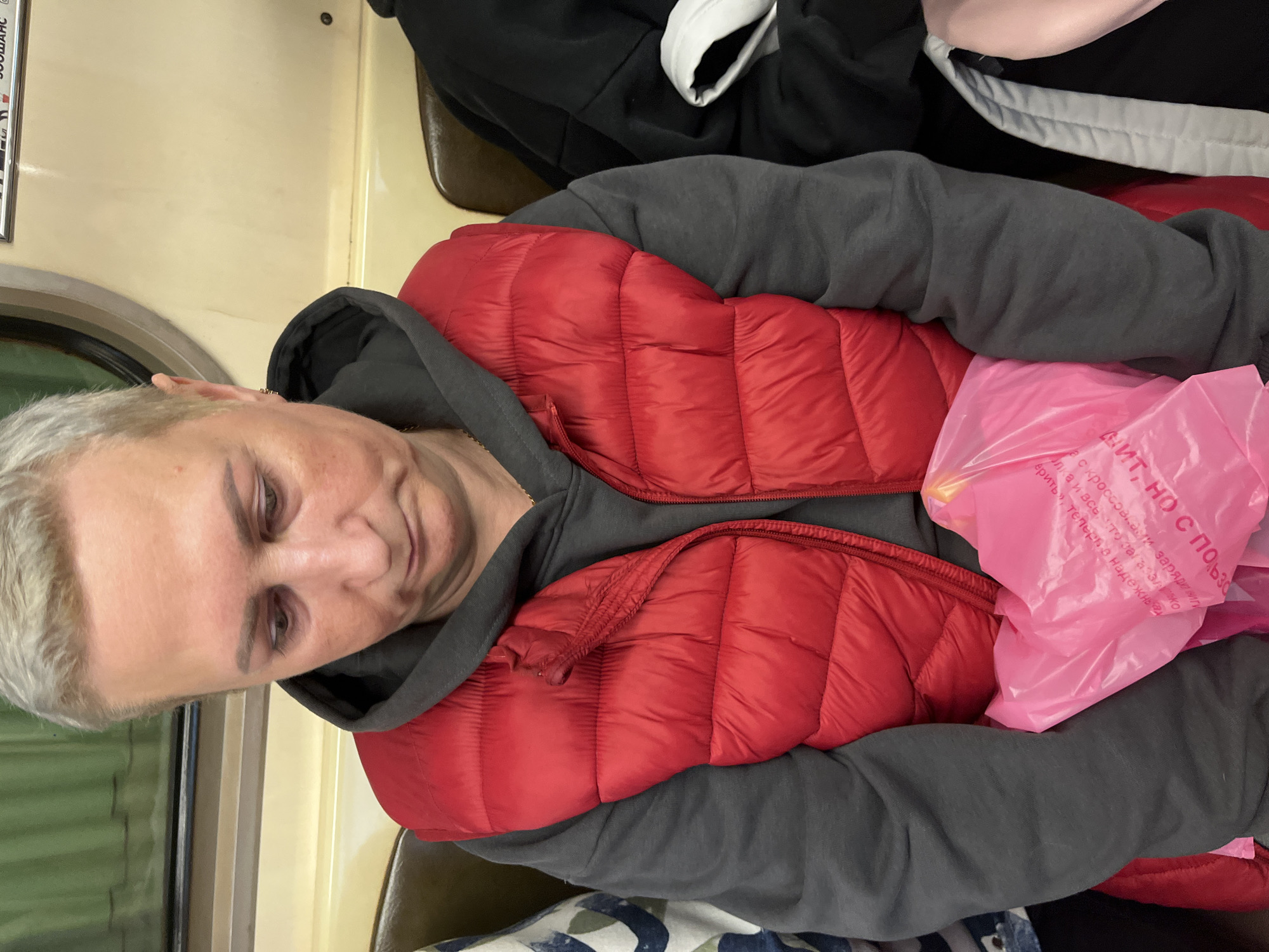 An adult individual with short, light-colored hair, wearing a dark gray hooded sweatshirt and a bright red quilted puffer vest, is seated in a metro train car in Minsk, Belarus. The person's gaze is directed downwards, suggesting a pensive demeanor. A small gold hoop earring is visible in the right ear, and a faint gold chain is around the neck. Resting in their lap is a bright pink plastic bag with partially visible Cyrillic text, including "ЗИНТ НО С ПОЛЬЗO" (ZINT, but with use), a brand associated with Belarus. To the right, another person is partially visible, wearing a dark garment with a white cuff. The interior of the train features light beige/cream-colored walls, light brown/tan upholstered seats with darker trim, and a window on the left showing a blurred green exterior, indicative of movement. A sign with Cyrillic text, including "ЗООШАНС" (ZOOSHANS), is partially visible on the upper right wall, further confirming the location in Minsk, Belarus.