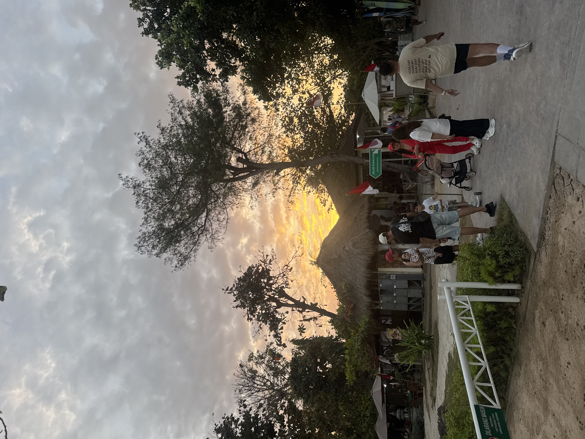 The image depicts a sunset scene at a beachside area in Kuta Selatan, Indonesia. The sky is filled with dramatic clouds tinged with orange and yellow hues from the setting sun. In the foreground, a white metal barrier with a green sign reading "DILARANG MASUK" (Forbidden Entry) and "JALUR EVAKUASI" (Evacuation Route) is visible.
Several people are present in the scene. On the left, a group of individuals stands near a thatched-roof structure, with umbrellas providing shade. A man in a black t-shirt and shorts is standing with a child in his arms, facing the camera. Beside him, a man in a black t-shirt with white lettering on the back and dark shorts is walking away from the camera. To the right of the man in the black t-shirt, a woman in a red jumpsuit and a white t-shirt stand with a baby stroller between them. Further to the right, a woman in a white t-shirt and black pants stands with her back to the camera. Another man in a beige t-shirt with text and dark shorts is walking away from the camera.
The environment includes lush green trees and vegetation surrounding the structures. Several Indonesian flags are displayed on poles. In the background, more thatched-roof buildings and an umbrella are visible. The ground surface appears to be a mixture of sand and paved walkways. The overall atmosphere suggests a public space, possibly a recreational area or dining establishment by the beach.