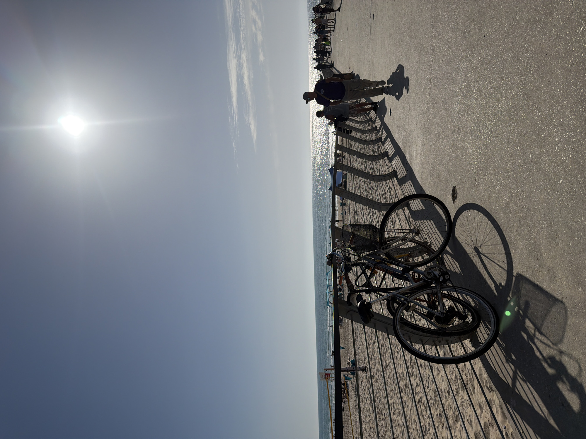 The image depicts a sunny scene at Bat Yam beach in Israel. The perspective is along a boardwalk or elevated walkway, where bicycles are parked along a railing. The beach and sea stretch along the horizon. People are visible on the boardwalk, casting shadows. The sky is bright, with the sun visible.