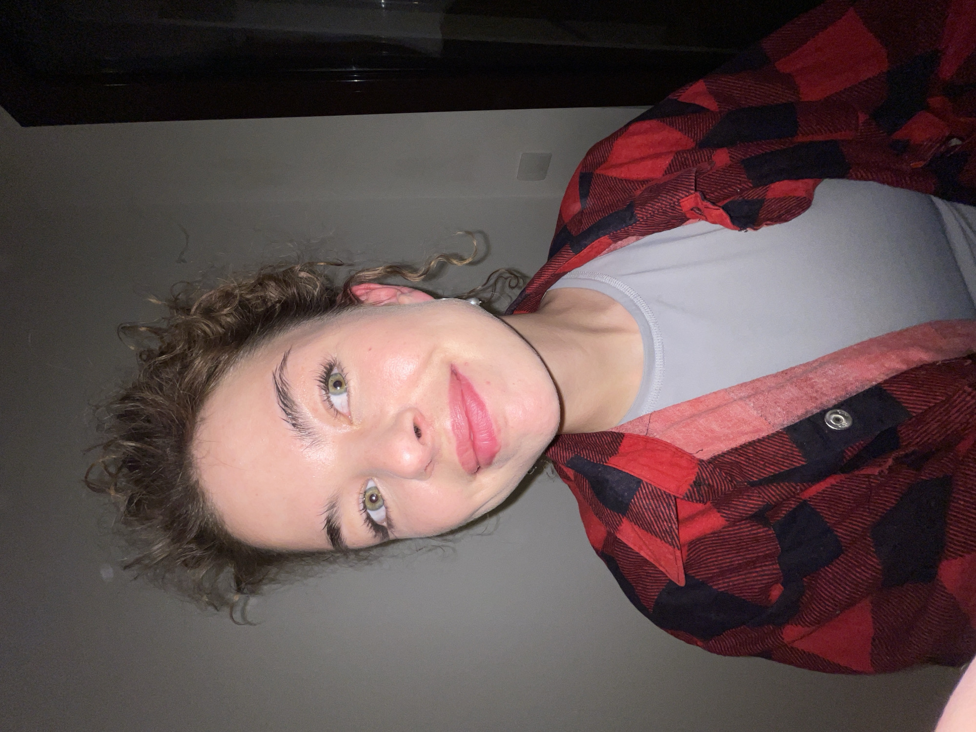 A person with fair skin, curly brown hair, and green eyes is looking at the camera. They are wearing a light gray t-shirt and a red and black plaid flannel shirt. The background is a gray wall.
Boreti, Montenegro is not identifiable from the image.